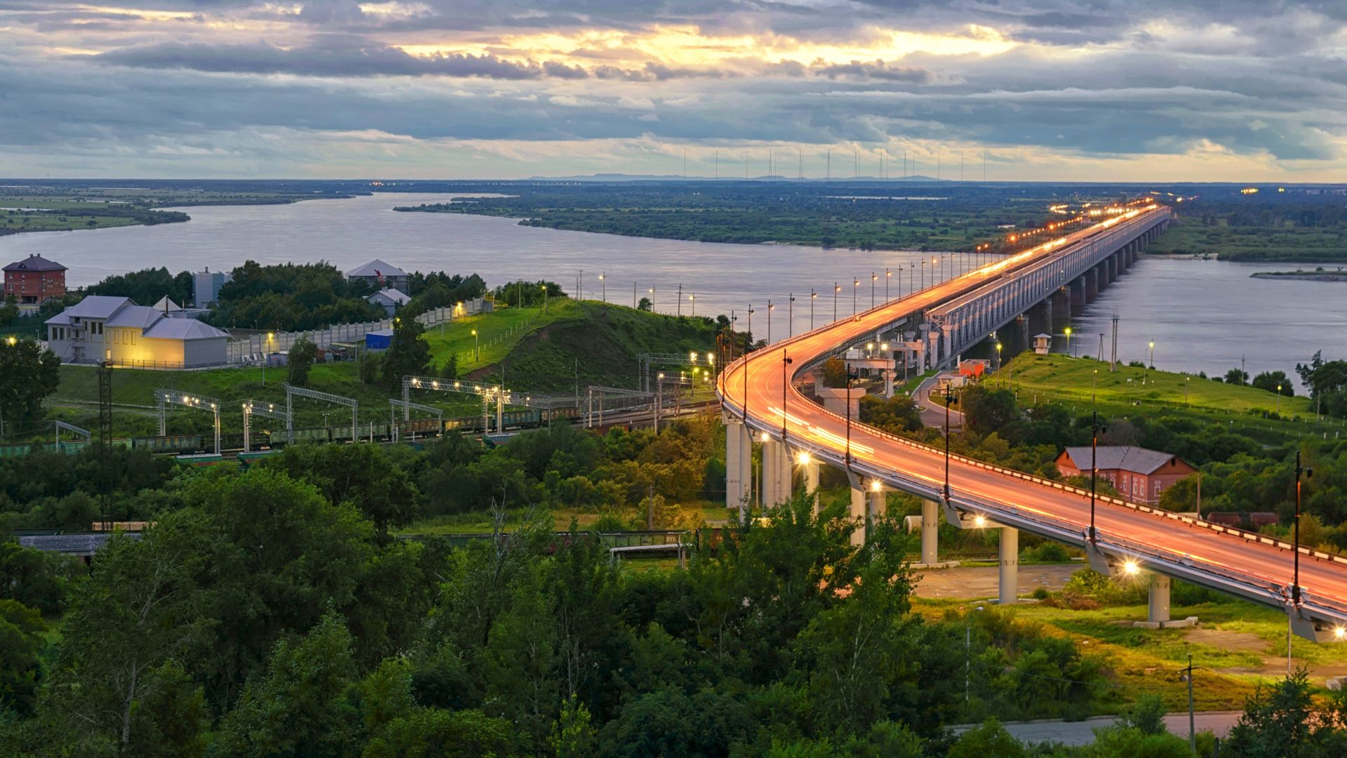 A long, illuminated bridge arches over a wide river at twilight or sunset. The photograph, taken from a high vantage point, showcases the Khabarovsk Bridge (Amur Bridge), with its curved roadway bright with lights against the dimming sky, lush green hills and a small settlement visible on the riverbanks.