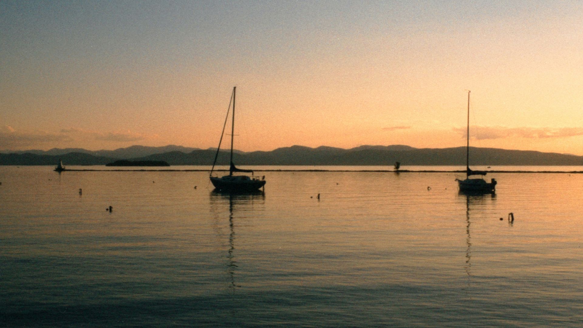 A scenic photograph capturing the silhouette of two sailboats anchored on the calm waters of a large lake during a beautiful orange and yellow sunset, with a distant, dark mountain range visible across the horizon.