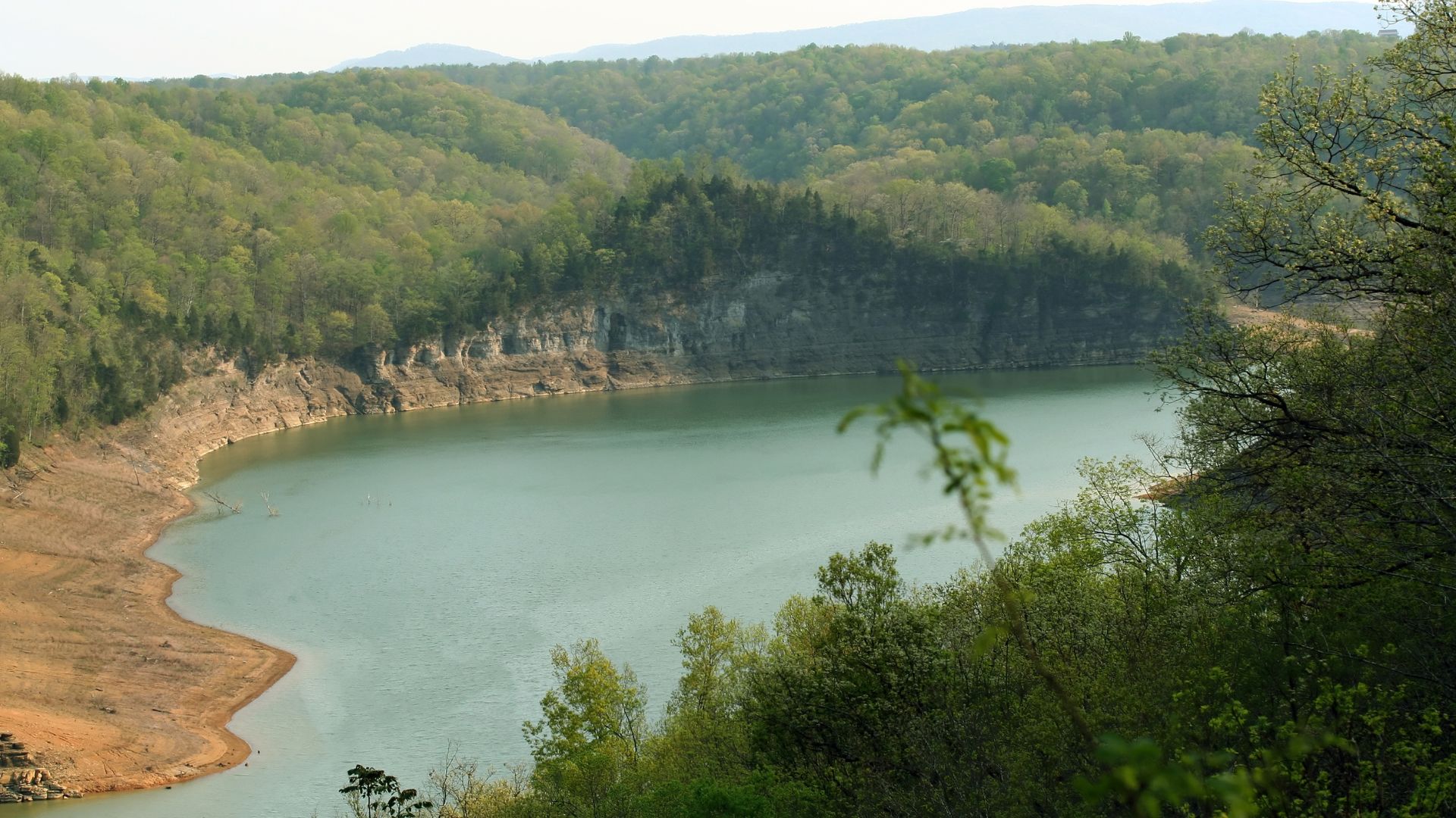 A scenic, elevated photograph of a large, tranquil lake bordered by steep hills covered in dense green forest, featuring an exposed, light-colored rocky and muddy shoreline due to low water levels.