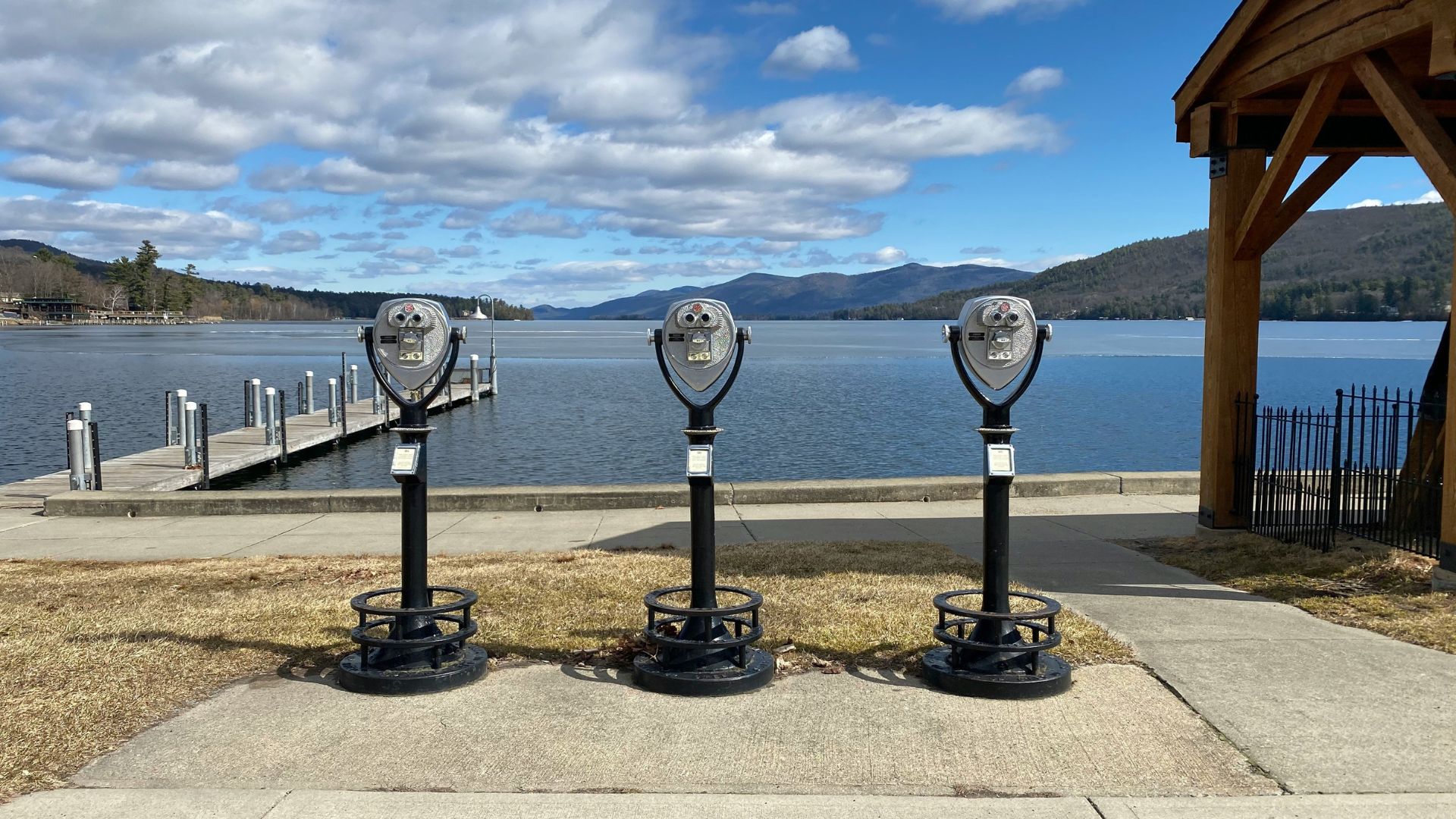 Three public, coin-operated binoculars on a concrete observation area overlook a wide blue lake with wooded mountains under a partly cloudy sky.