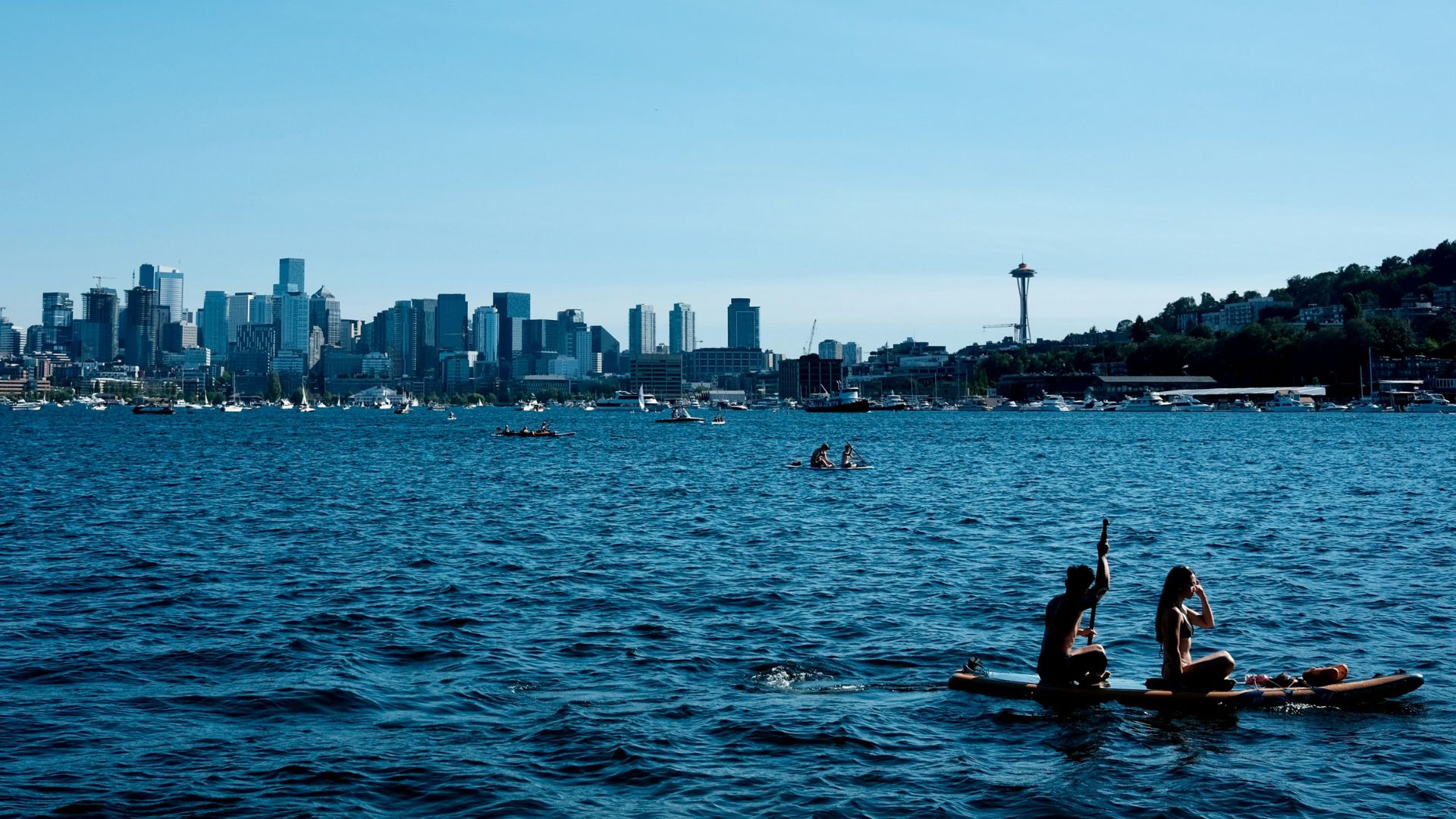A wide, sunlit view across the dark blue water of Lake Union, with two people on a stand-up paddleboard in the foreground and the Seattle skyline, including the iconic Space Needle, visible in the distance against a clear blue sky.