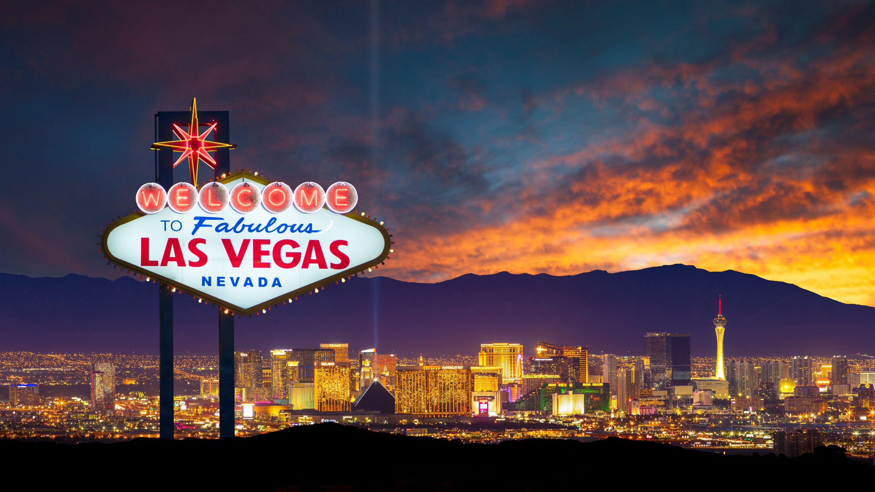 A night-time, wide view of the illuminated, Googie-style "Welcome to Fabulous Las Vegas Nevada" sign with the bright lights of the Las Vegas Strip and mountains visible in the background under an orange and dark sky.