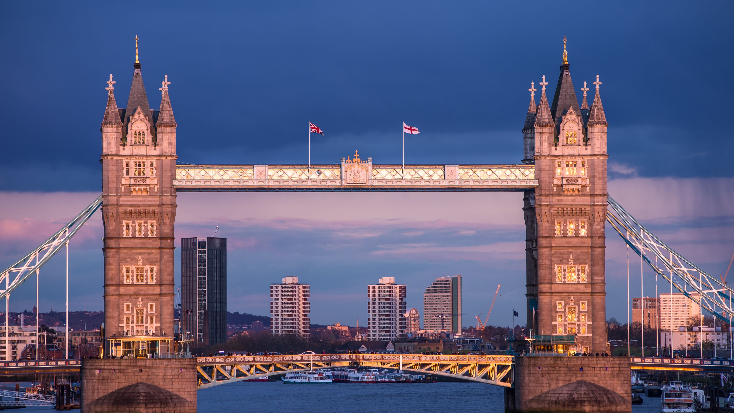 A wide-angle view of the illuminated, Neo-Gothic style Tower Bridge over the River Thames at twilight, with the city skyline including several modern skyscrapers visible in the background.