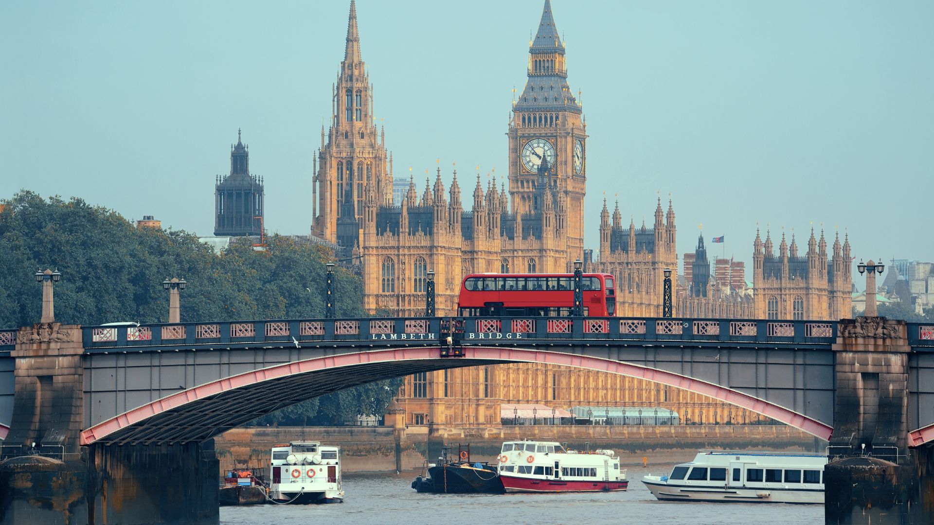 The iconic Elizabeth Tower (Big Ben) and the Palace of Westminster are visible across the River Thames, with a red double-decker bus crossing a bridge in the foreground.