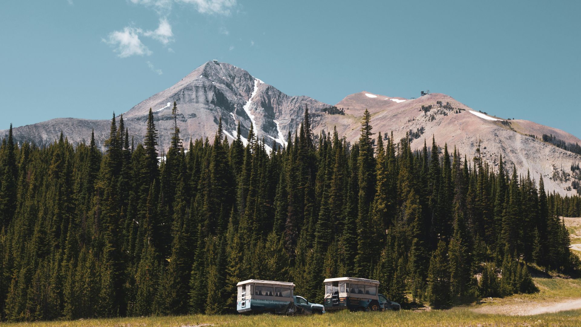 A landscape photograph of a rugged, snow-dusted mountain peak rising above a dense evergreen forest. In the grassy foreground, two old, slightly dilapidated buses or campers sit in a field under a bright blue sky with a single white cloud.