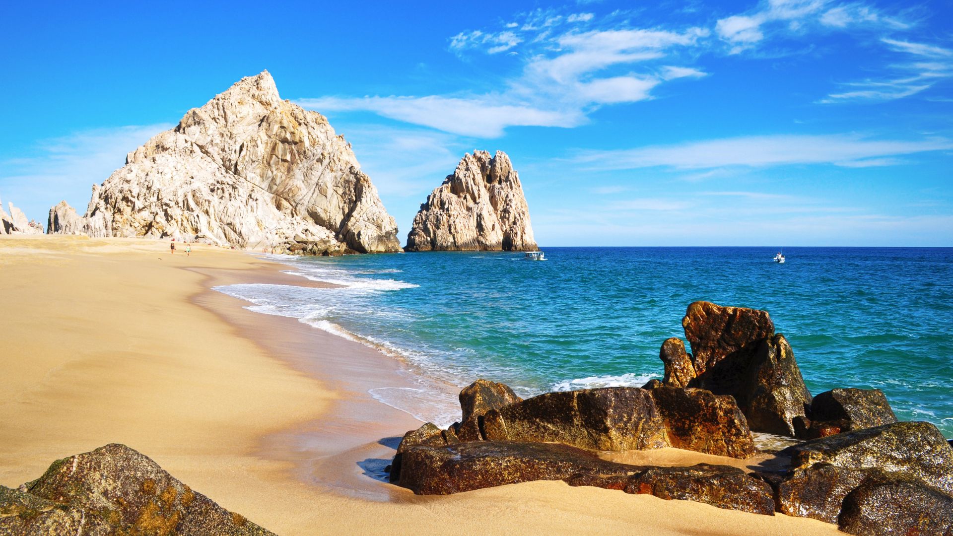 A bright, sunny view of the sandy Lovers' Beach in Cabo San Lucas, Mexico, with large, white, rugged rock formations rising from the sand and the vibrant turquoise ocean water. Darker rocks are in the immediate foreground, and a small white boat is visible in the distance.