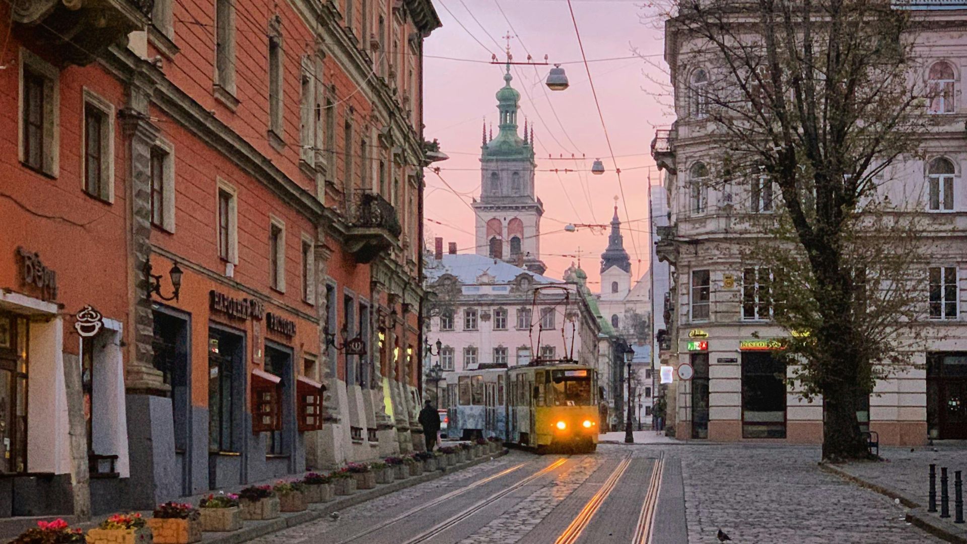 A vibrant evening photo of a cobblestone street in Lviv, Ukraine, where a yellow tram with illuminated headlights approaches. Historic, colorful European buildings line the road, with a church spire visible in the distance against a pink and purple twilight sky.