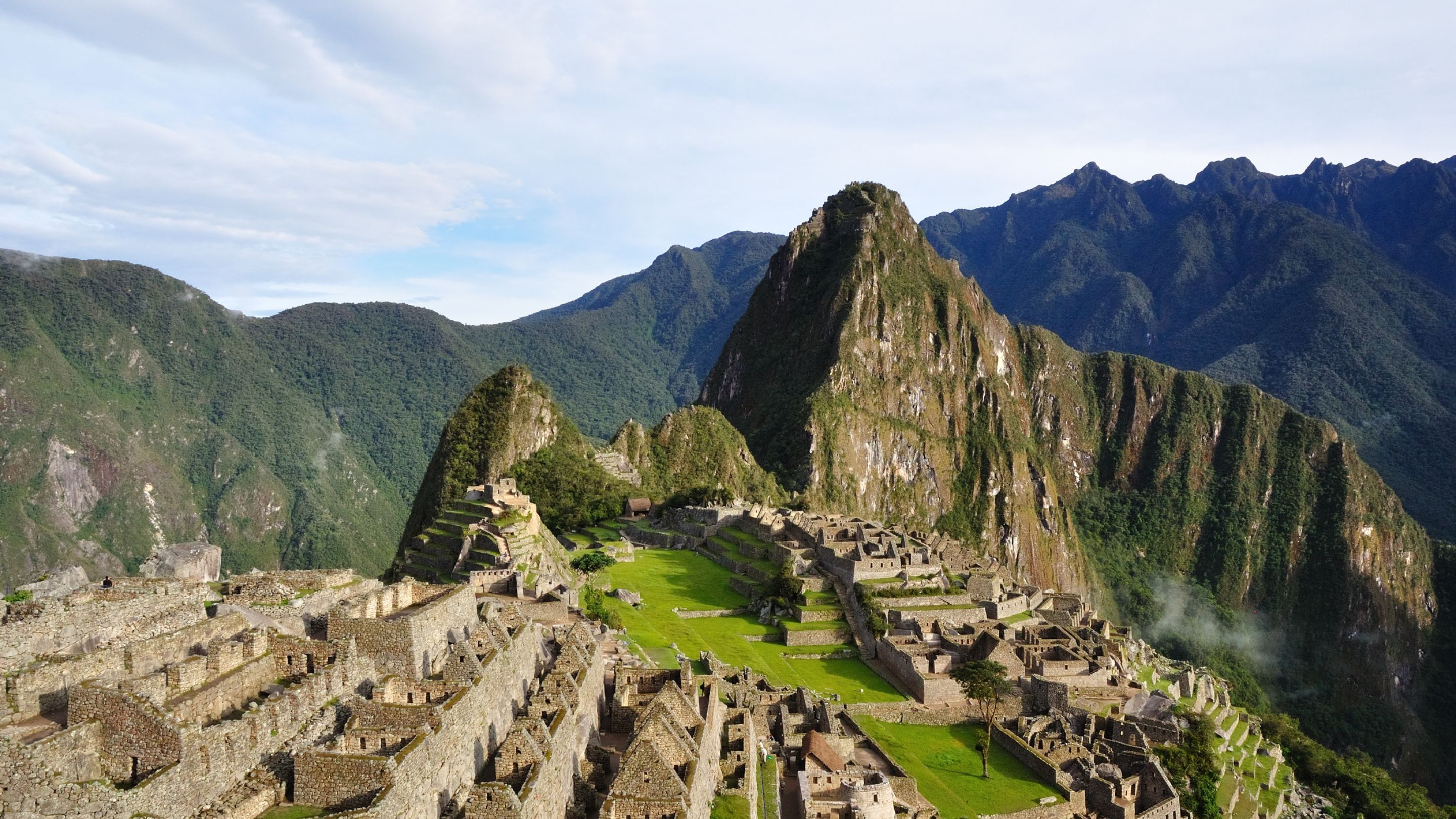 A wide, elevated view of the ancient, stone Inca ruins of Machu Picchu nestled amidst lush, green mountains under a partly cloudy sky, with the distinct peak of Huayna Picchu towering in the background.