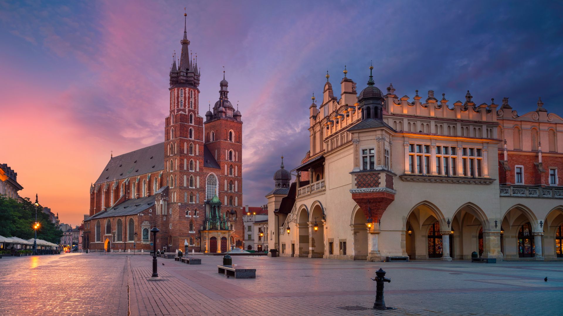 A wide-angle view of the historic Main Square in Kraków, Poland, at sunrise or sunset, featuring the tall Gothic towers of St. Mary's Basilica on the left and the long Renaissance-style Cloth Hall on the right, all under a dramatic sky of pink and blue.