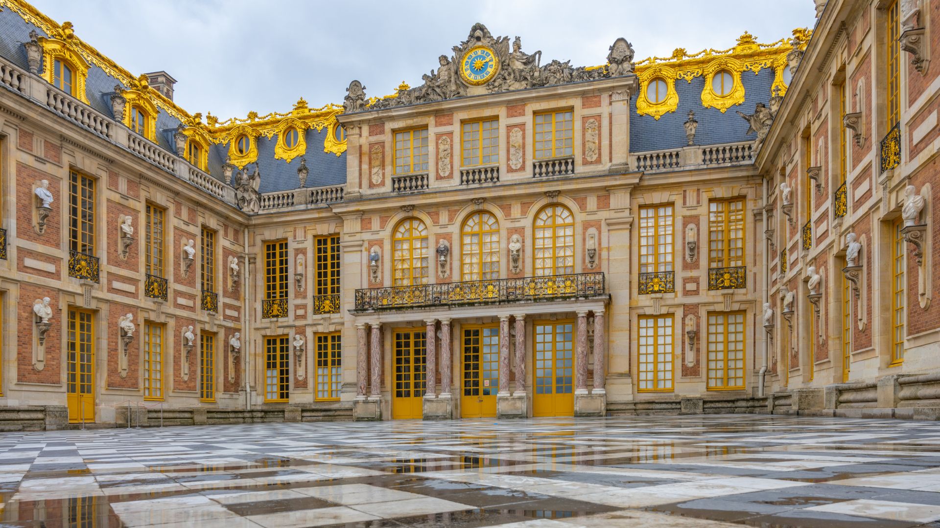 The historic Marble Courtyard of the Palace of Versailles, featuring a black and white checkered floor and an ornate brick and stone facade with extensive gold detailing.