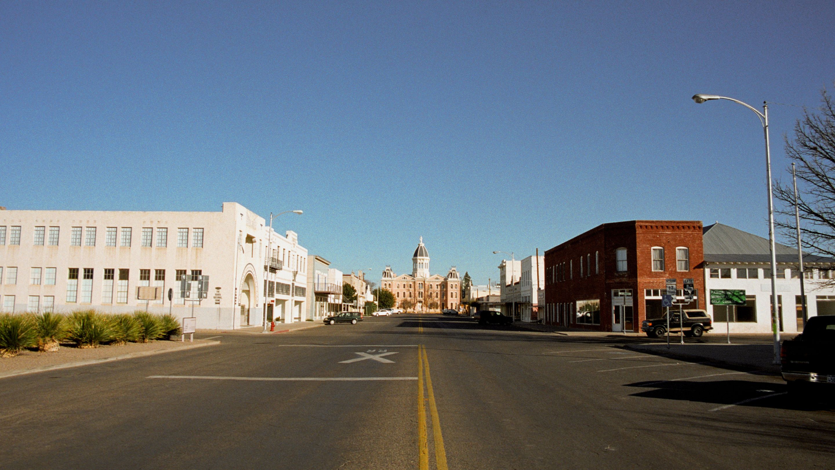 A wide-angle street-level view looking north up Highland Avenue in Marfa, Texas, towards the historic pink stucco Presidio County Courthouse building with its central dome and Lady Justice statue at the top.