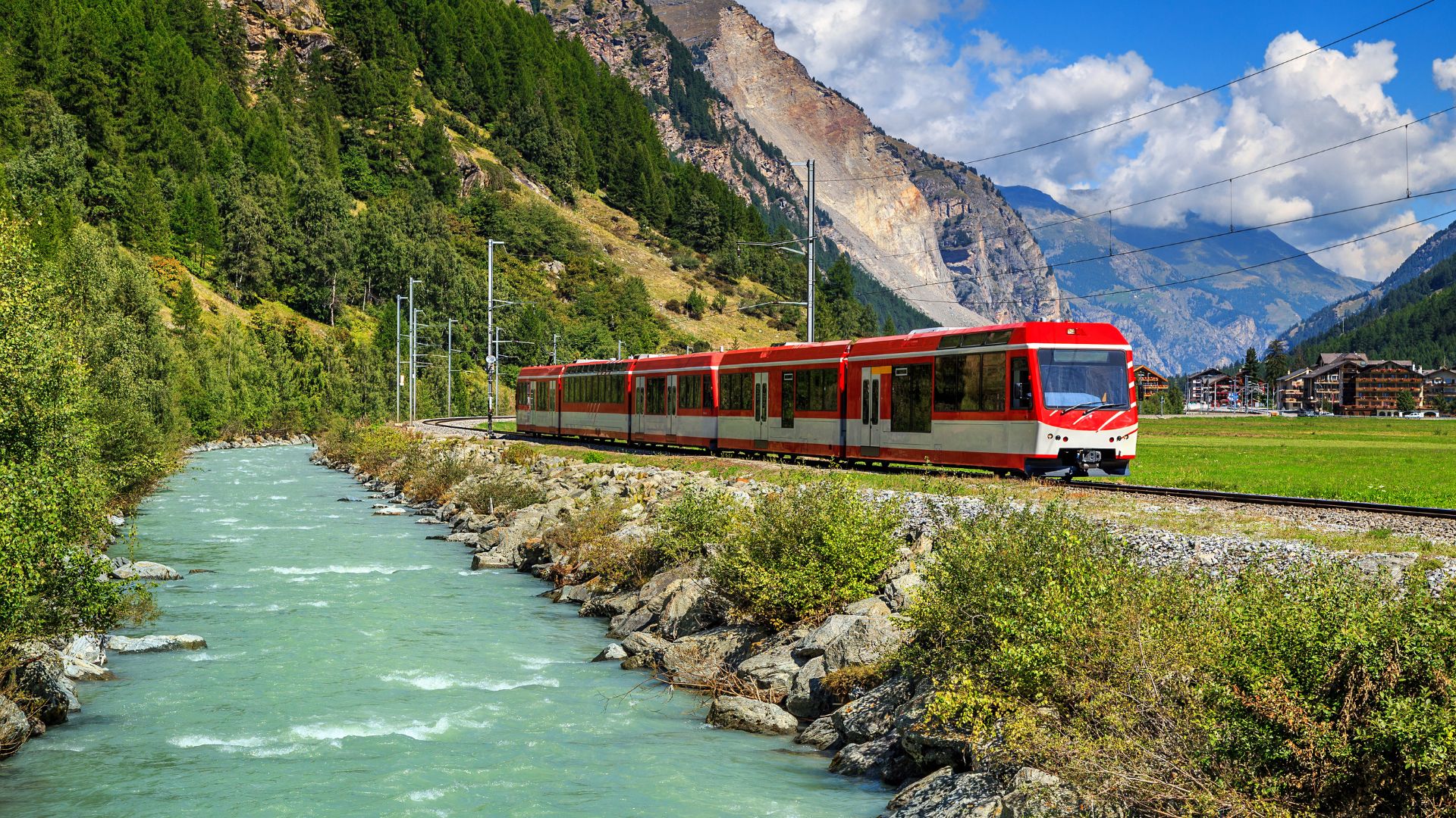A bright daytime photograph of a red and white electric multiple unit train operated by the Matterhorn Gotthard Bahn in a wide Swiss Alpine valley. The train is traveling along tracks running parallel to a vibrant, milky-turquoise river. The valley floor is lush green meadow, leading up to steep, forested mountain slopes and rugged gray peaks in the distance, with a clear blue sky above.