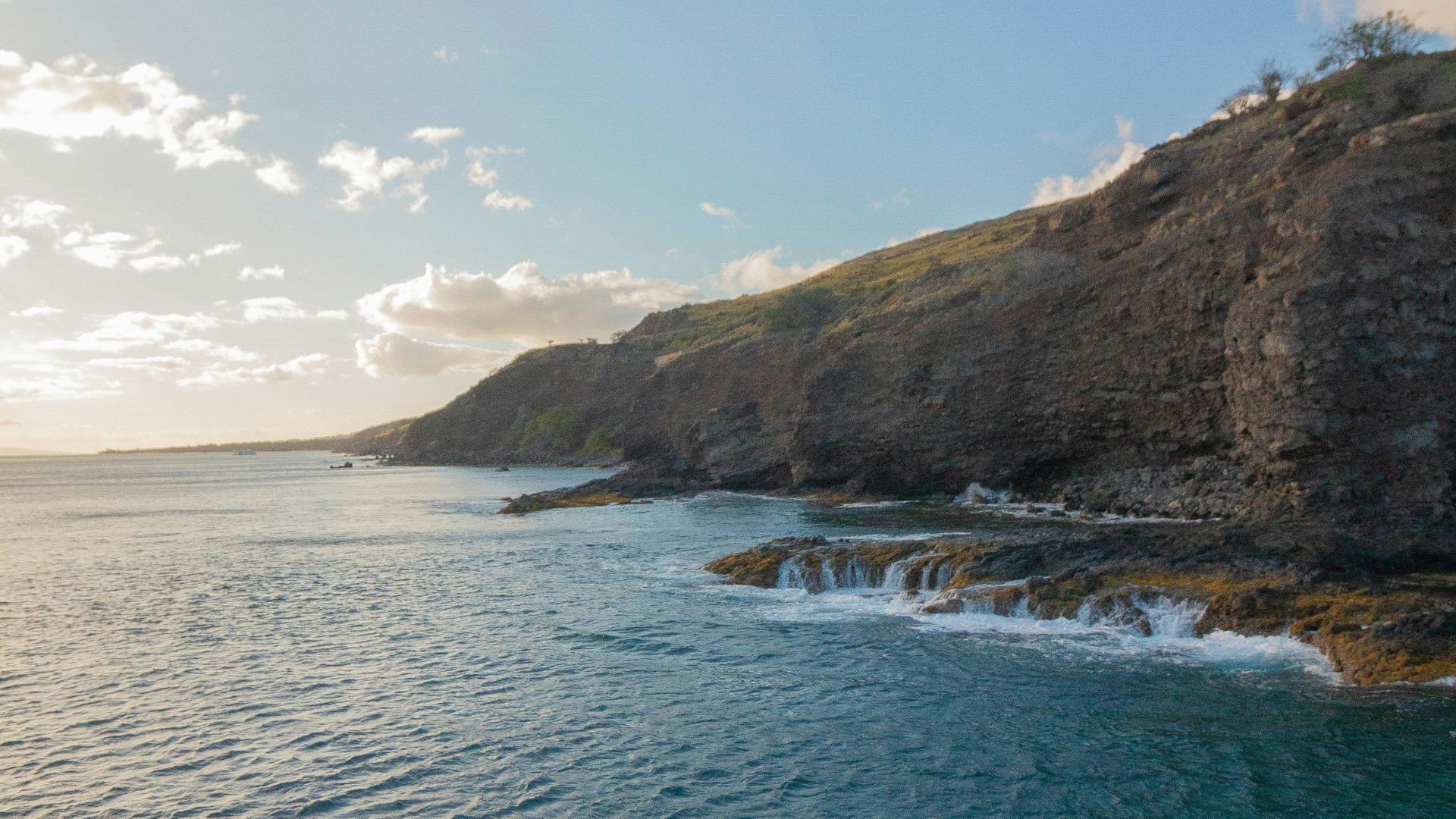 A sunlit photograph of the deep blue ocean water contrasting with a steep, rugged coastline of dark volcanic rock under a bright blue sky with scattered white clouds.
