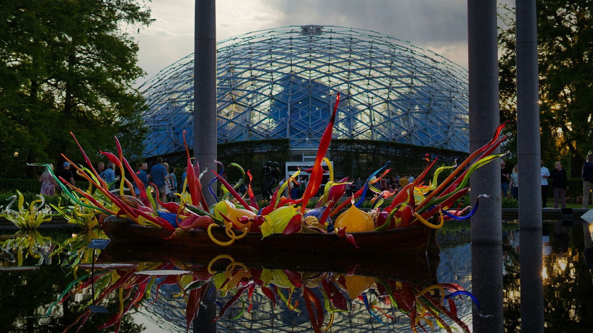 A photograph of the iconic geodesic-dome Climatron greenhouse at the Missouri Botanical Garden, with a wooden boat sculpture filled with colorful glass art floating in the reflecting pool during a Chihuly art exhibition.