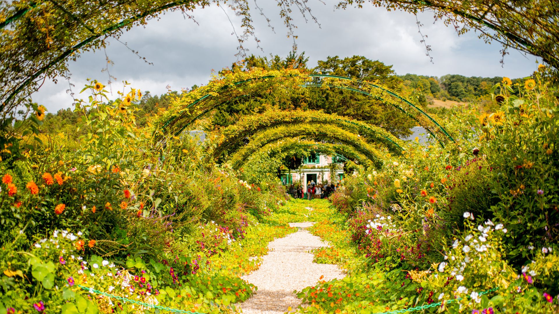 The iconic flower-lined central pathway with archways at Claude Monet's house and gardens in Giverny, France, under a partly cloudy sky.
