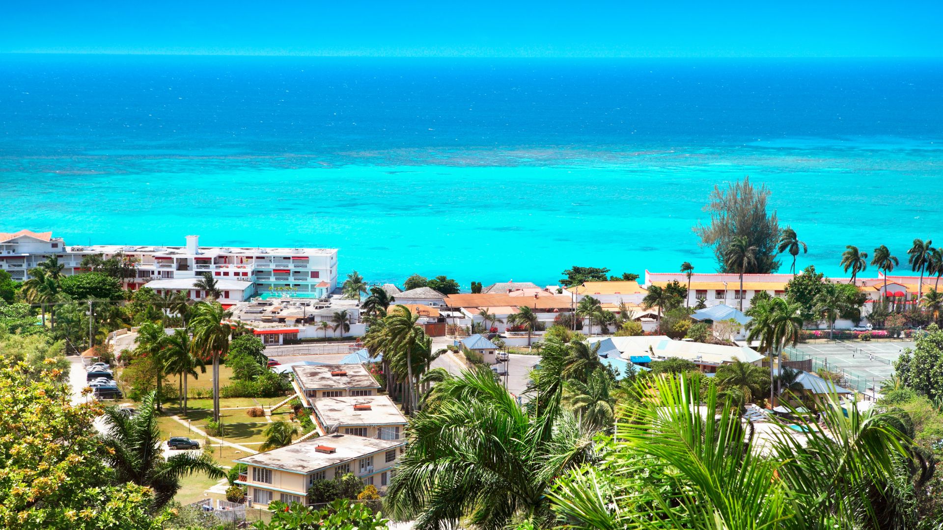 An elevated, panoramic view of the coastal town of Montego Bay, Jamaica, situated along the Caribbean Sea. The scene features various white and multi-colored buildings, lush green palm trees, and striking turquoise ocean water under a bright blue sky.