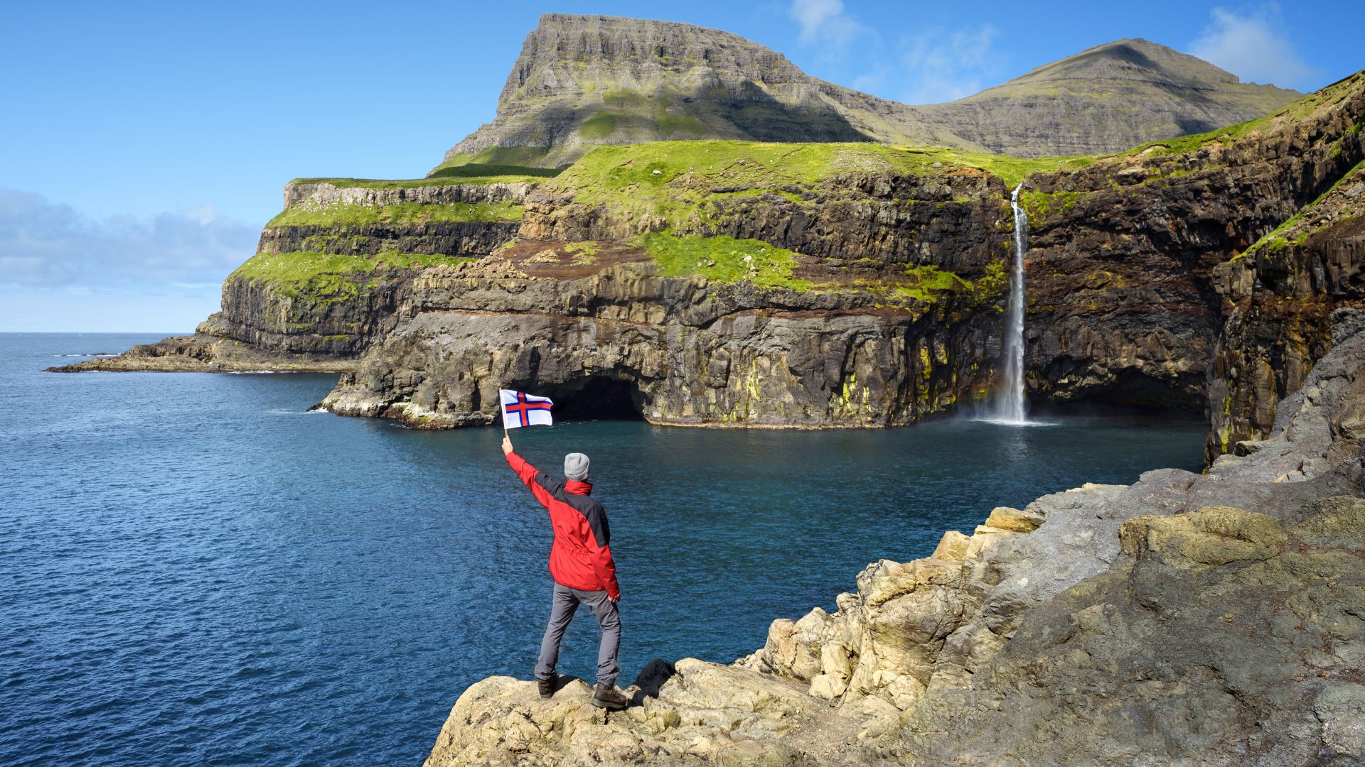 A daytime photograph of a person in a red jacket holding a flag, standing on a rocky cliff edge overlooking the dramatic Múlafossur waterfall as it plunges from a green cliff face directly into the deep blue North Atlantic Ocean.