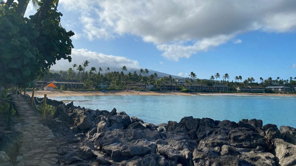 A path made of large dark gray rocks leads the viewer toward a crescent-shaped, light sand beach and calm, turquoise bay water, lined with palm trees and resort buildings, all under a bright blue sky with large white clouds.