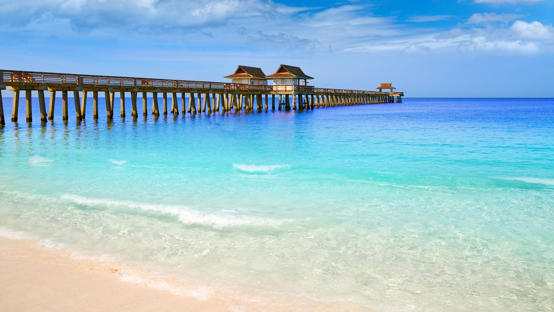 A long, wooden pier with covered gazebos extends far into the calm, turquoise waters of the Gulf of Mexico, viewed from a white sandy beach with gentle waves under a bright blue sky with white clouds.