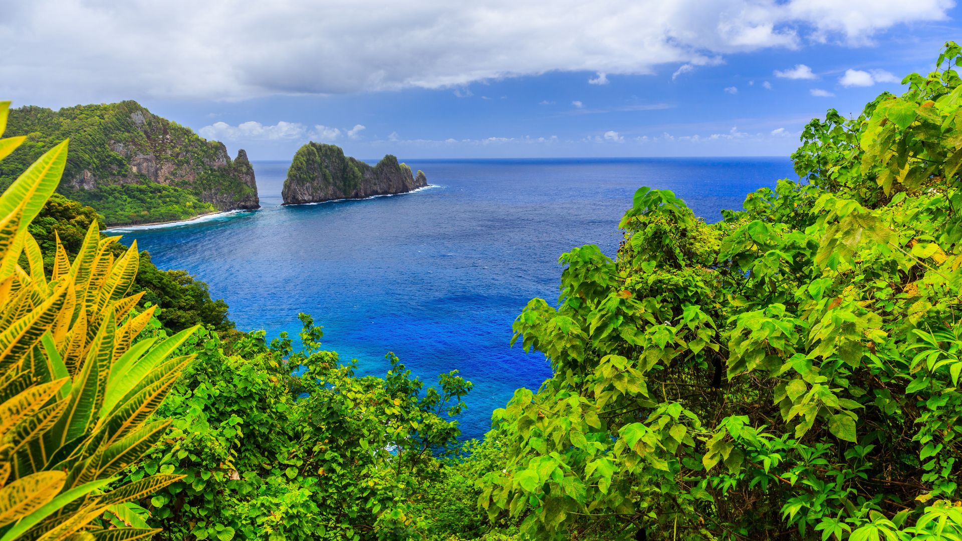 An elevated, panoramic view of a lush green tropical island coastline with steep cliffs and rocky islets in a bright blue ocean under a partly cloudy sky.