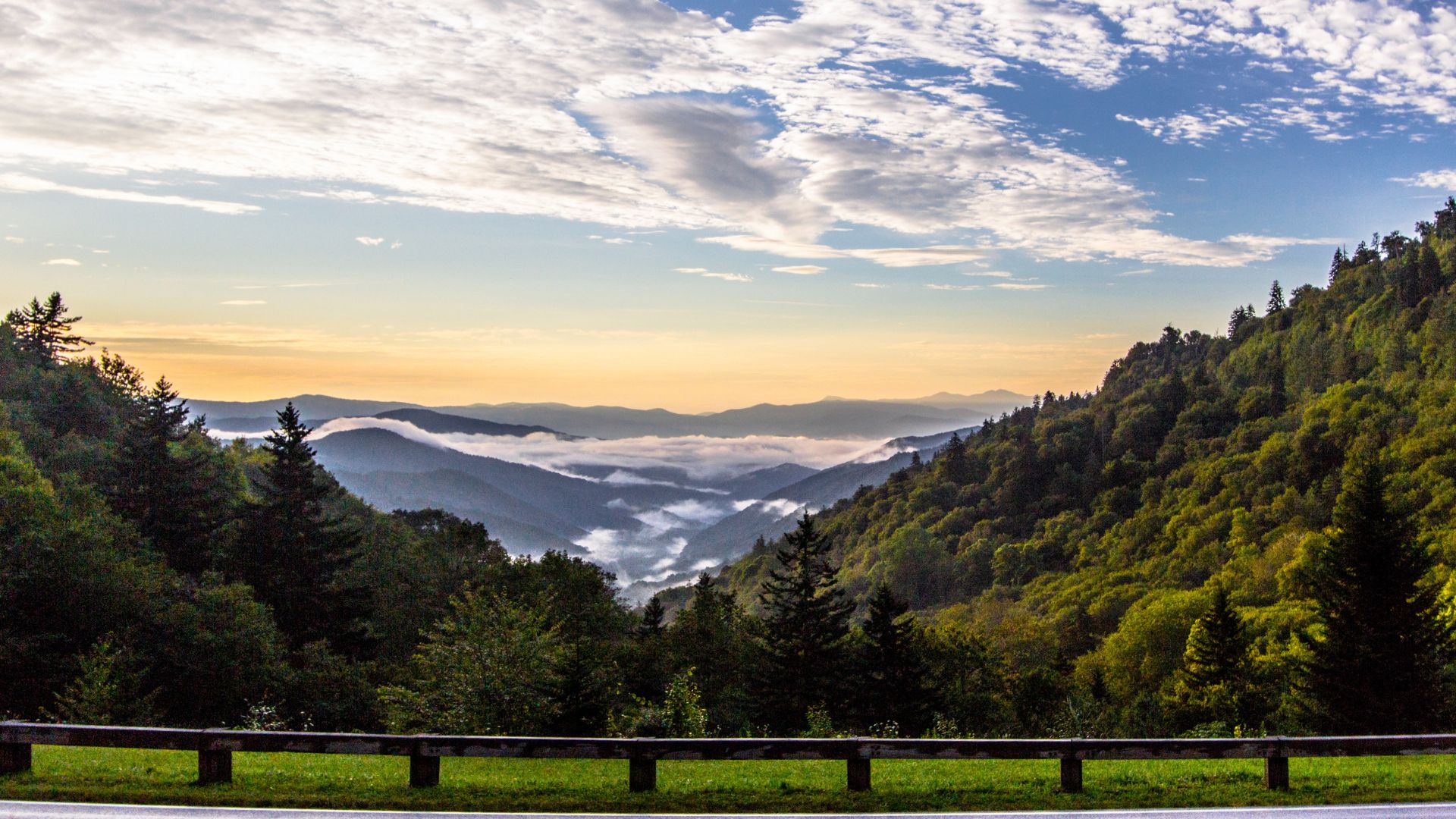 A mountain landscape view at sunrise showing layers of tree-covered ridges disappearing into a misty valley, as seen from an overlook with a guardrail in the foreground.