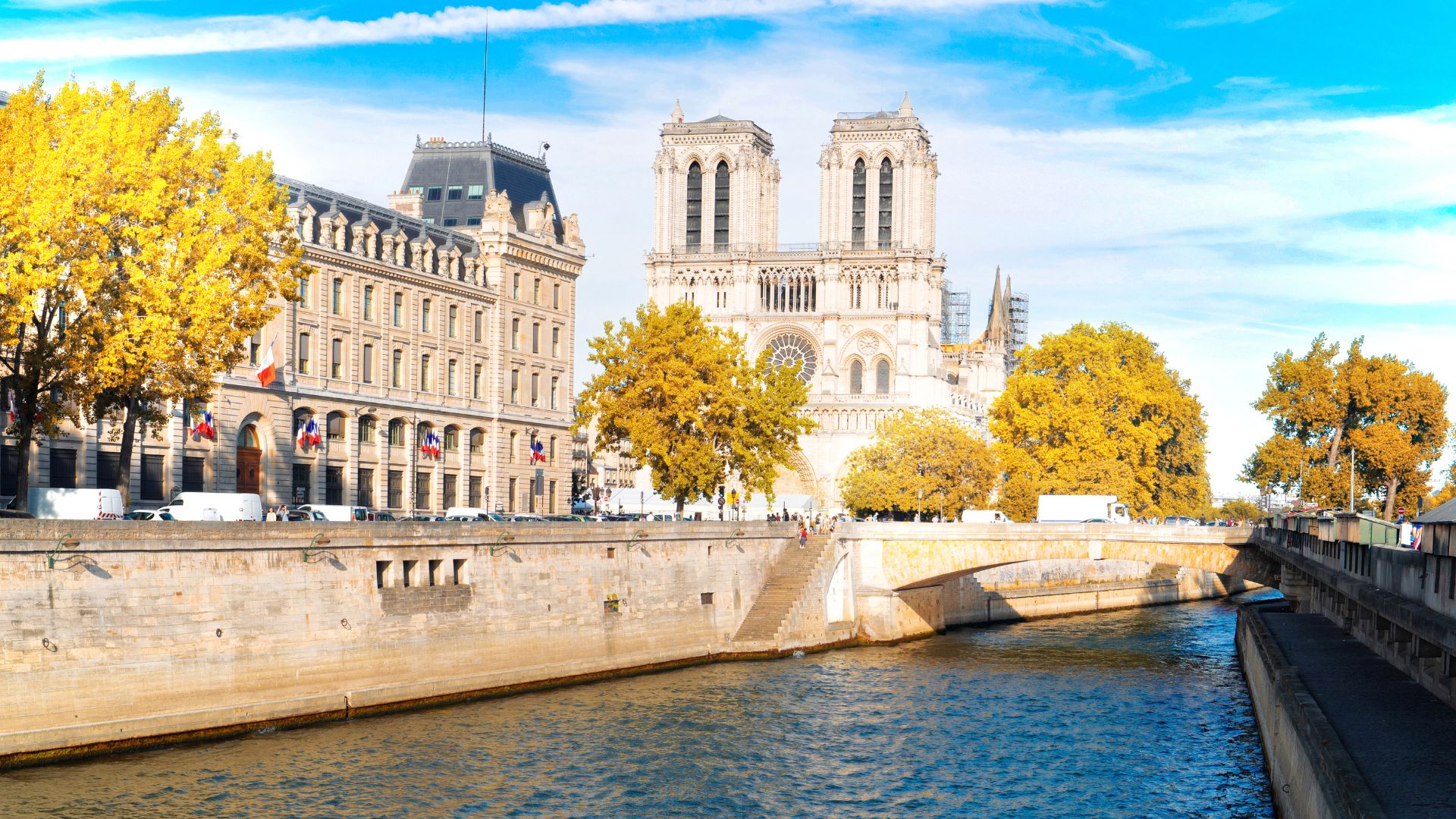 A view across the Seine River of the historic Gothic architecture of Notre-Dame Cathedral and an adjacent building on the Île de la Cité, surrounded by autumn trees under a blue sky.