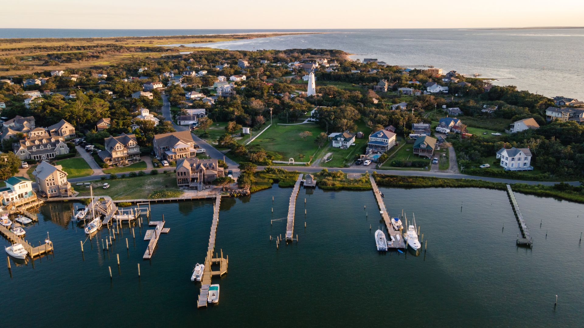 An aerial view of the small, coastal village of Ocracoke, North Carolina, showing houses among lush green trees, wooden docks and boats on the harbor in the foreground, and a white lighthouse visible in the center, with a narrow strip of land separating the harbor from the vast ocean in the background.
