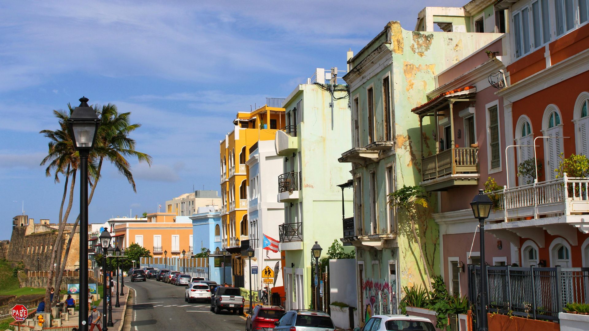A bright, daytime photo of a narrow street in Old San Juan, Puerto Rico, lined with tall, colorful Spanish colonial buildings featuring balconies and a few parked cars. Palm trees line the left side of the street under a blue sky.