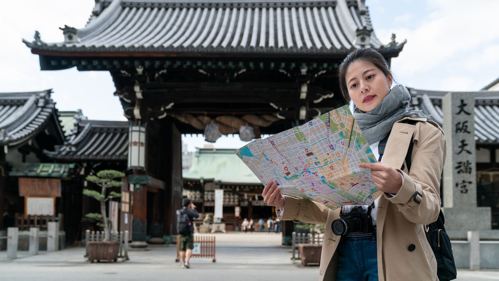 A female tourist with a map in the courtyard of the Osaka Tenmangu Shrine in Japan, with the large entrance gate visible in the background.