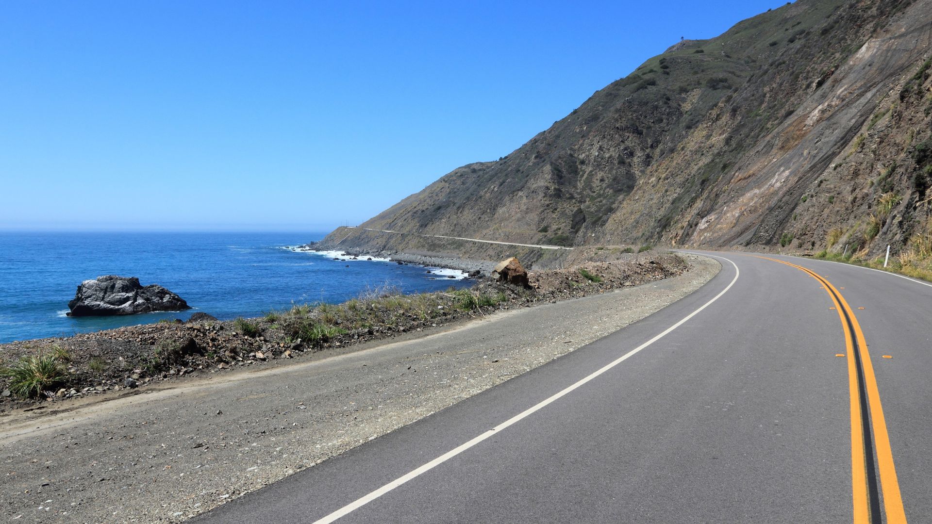 A scenic view of a paved coastal highway, California State Route 1, curving alongside a steep mountain cliff above the deep blue ocean under a clear sky. A large rock formation is visible offshore.