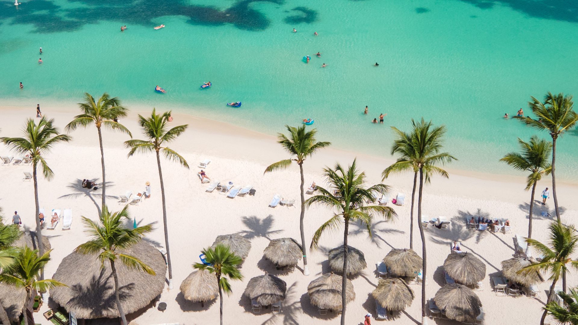 An aerial view of a crowded tropical beach with white sand and vibrant turquoise ocean water. Numerous palm trees and thatched sun umbrellas line the shore, with several people swimming and enjoying water activities in the sea.