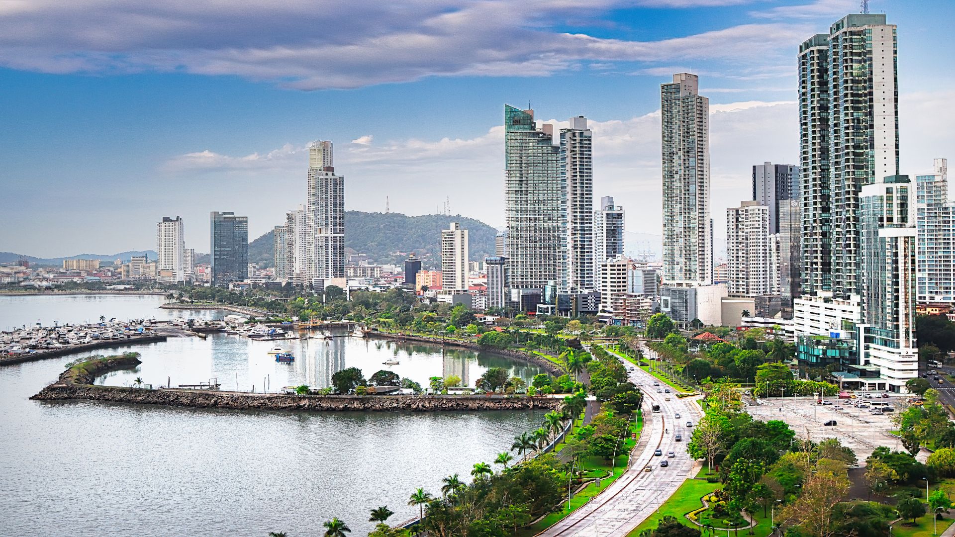 Daylight panoramic view of the modern, high-rise skyline of Panama City, Panama, lining a bay with a busy coastal highway and a small marina.