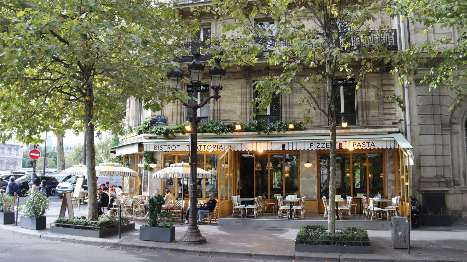 A charming Parisian street view of the "Bistrot Trattoria" restaurant with outdoor seating, striped awnings, and large trees lining the sidewalk.