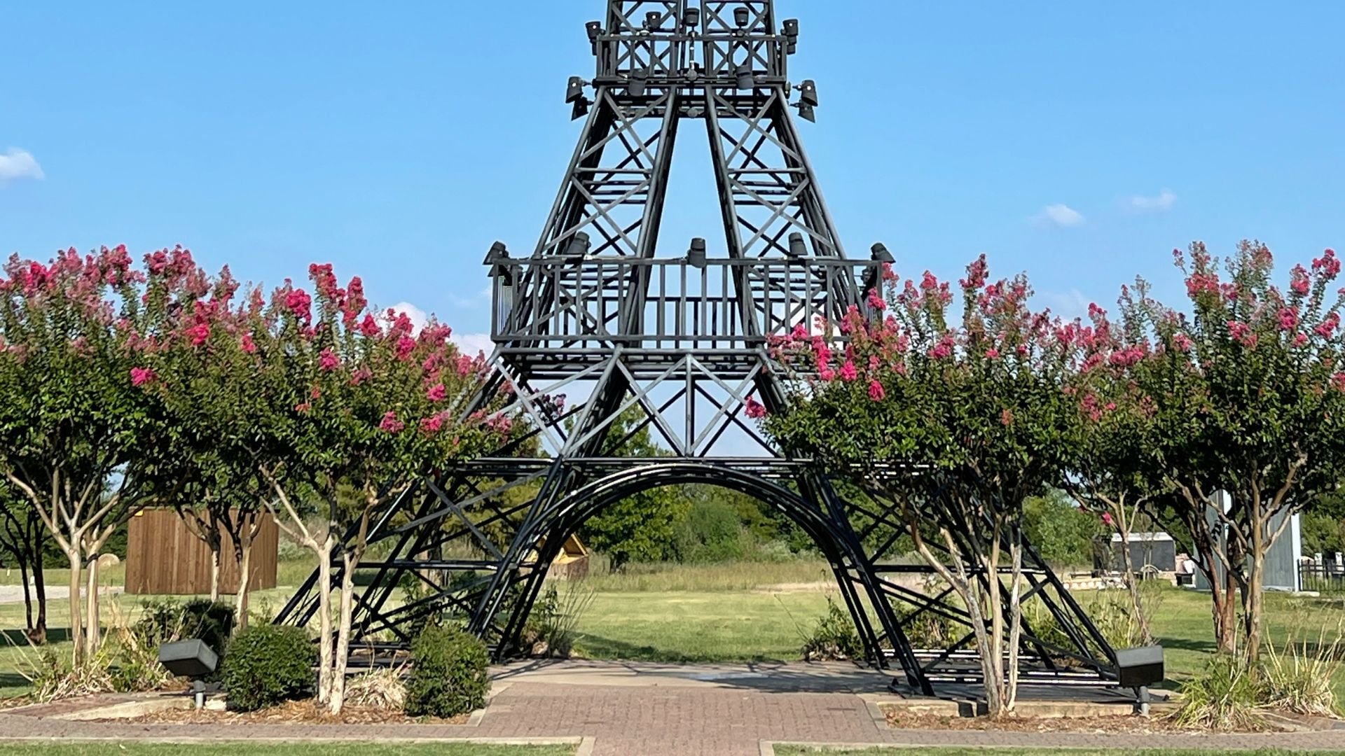 A 65-foot-tall, black steel replica of the Eiffel Tower is topped with a large, bright red cowboy hat, set against a blue sky with pink flowers in the foreground.