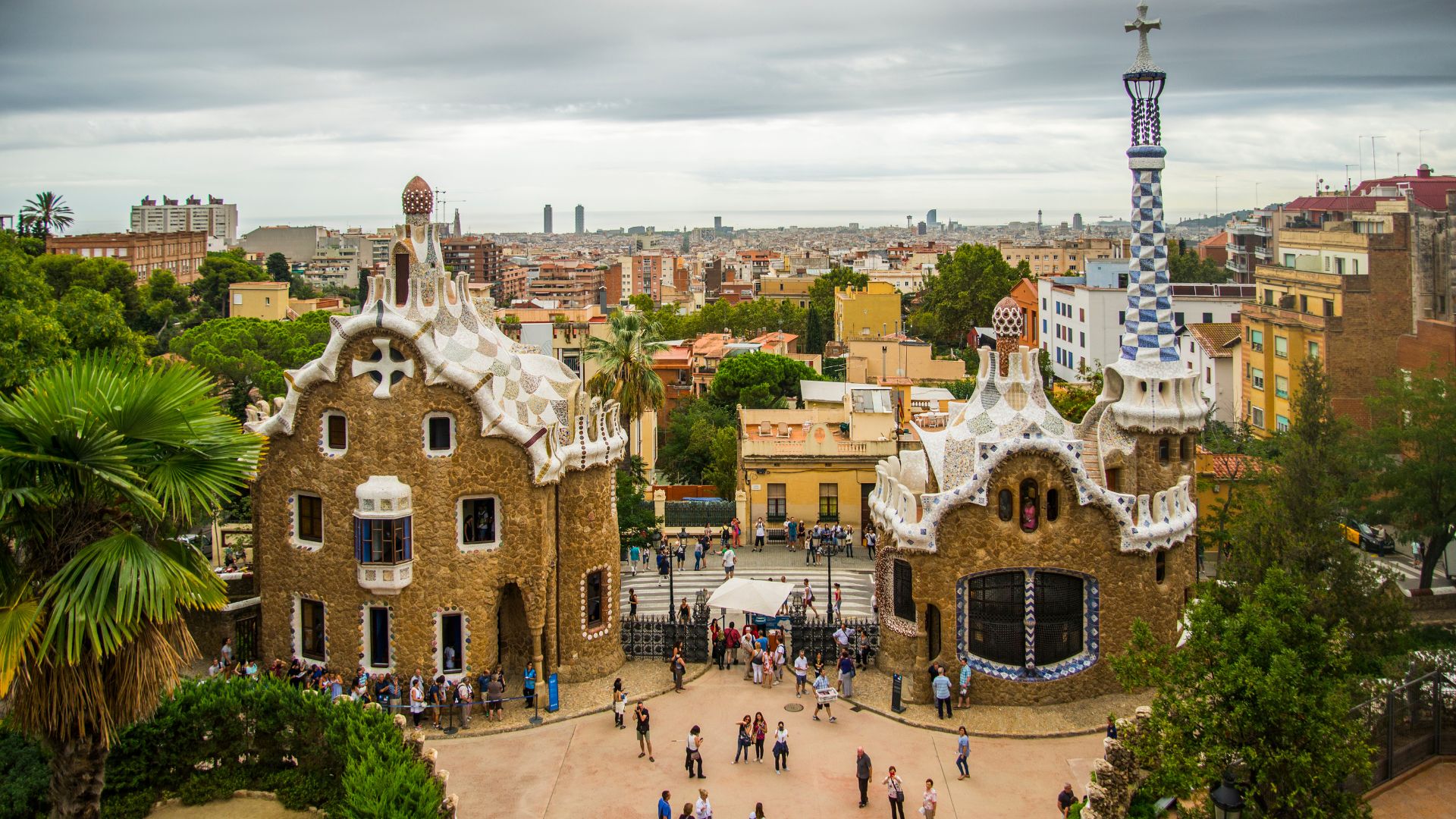 An elevated view shows two fantastical, gingerbread-style stone buildings with curved, "trencadís" mosaic-covered roofs, one featuring a tall, checkered spire with a cross on top. Below them, a plaza filled with tourists leads to the main entrance of Park Güell, with the sprawling city of Barcelona visible in the background under a cloudy sky.