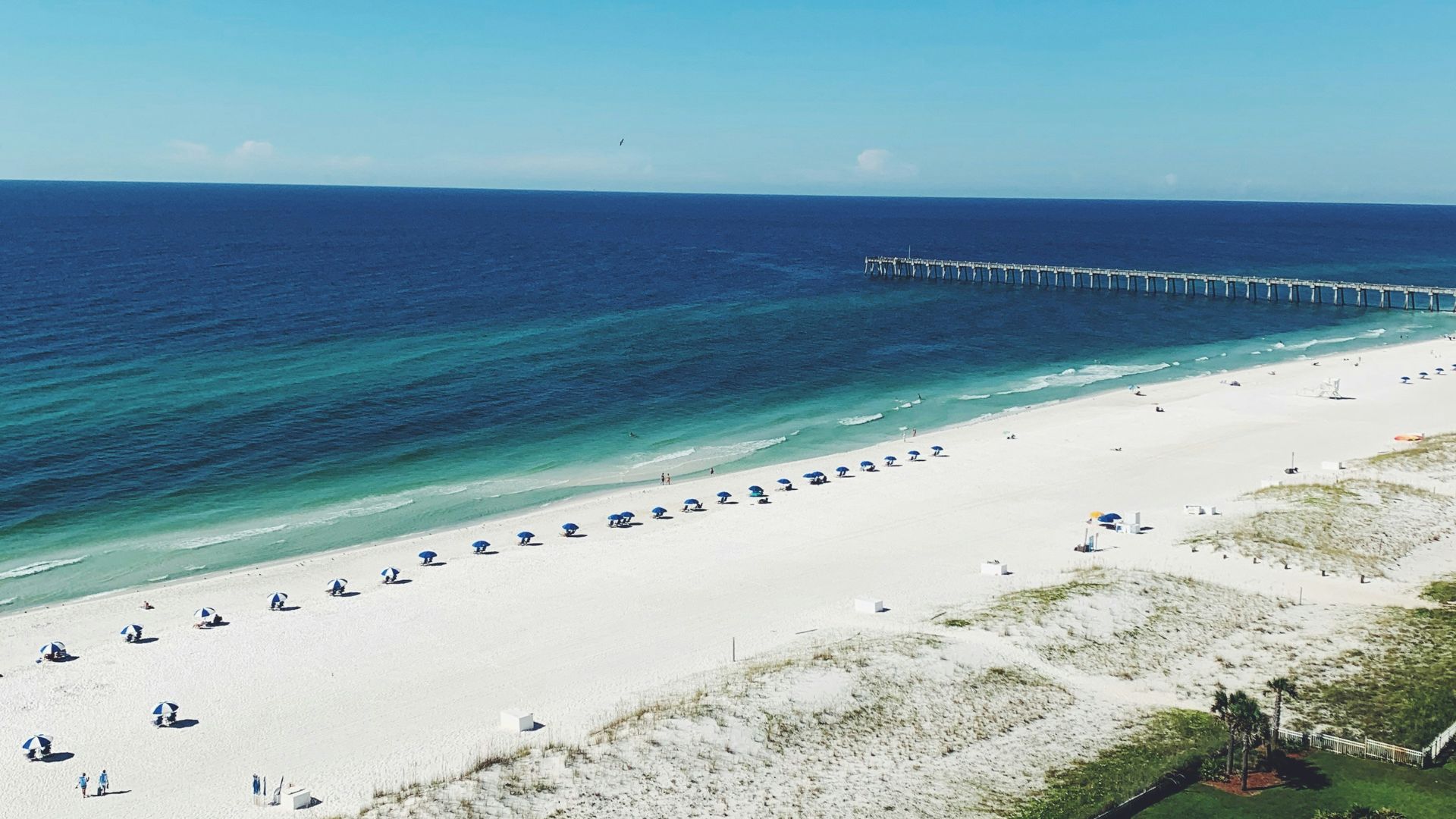 An aerial photograph of the long, white-sand Pensacola Beach in Florida, showing the long fishing pier extending into the bright turquoise Gulf of Mexico waters under a clear blue sky.