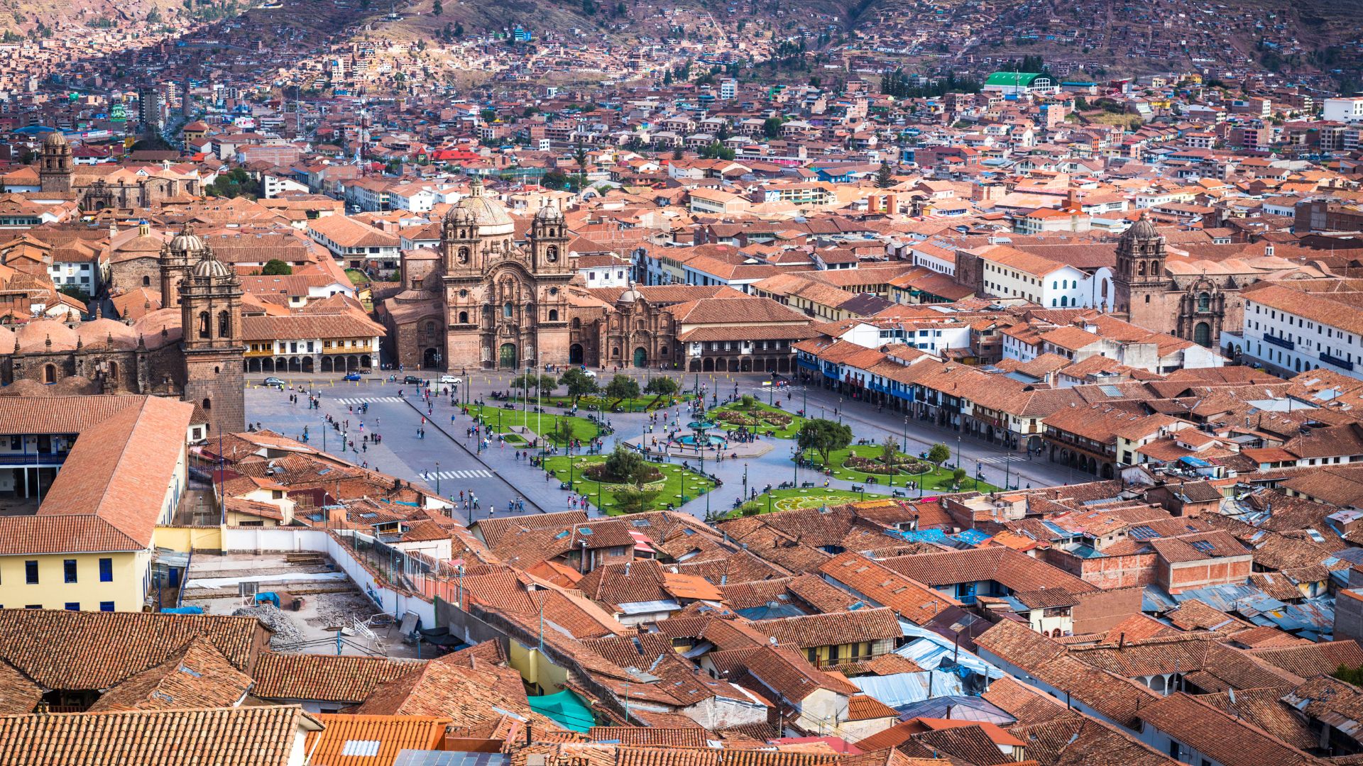 An elevated view of a large, busy city plaza surrounded by Spanish colonial buildings with red-tiled roofs, nestled in a valley with steep, green mountains rising in the background.