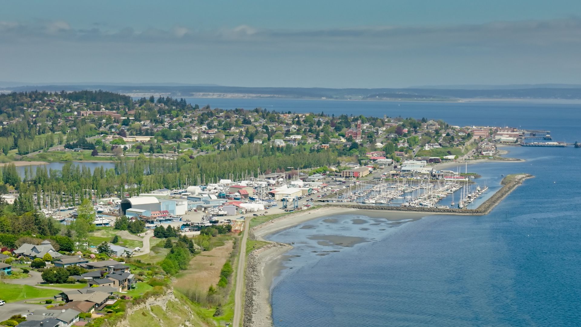 An aerial panoramic view of a coastal town with a large marina filled with docked boats, buildings lining the waterfront, dense green trees covering the hills, and a wide body of water in the background.