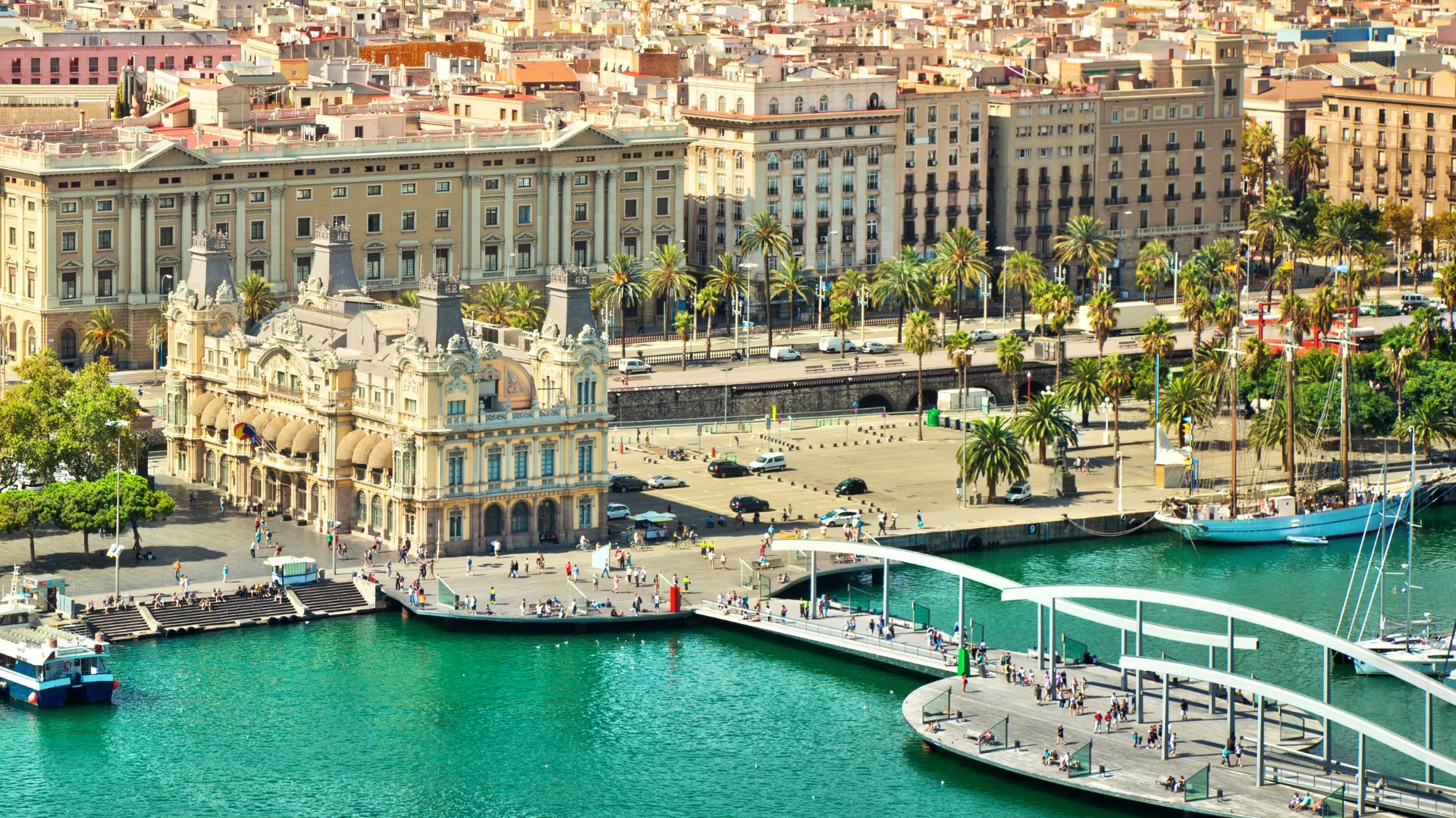 An elevated view of the busy Port Vell harbor in Barcelona, Spain, featuring historic buildings with classic facades, an avenue lined with palm trees, a modern white footbridge, and several boats docked in the turquoise water.