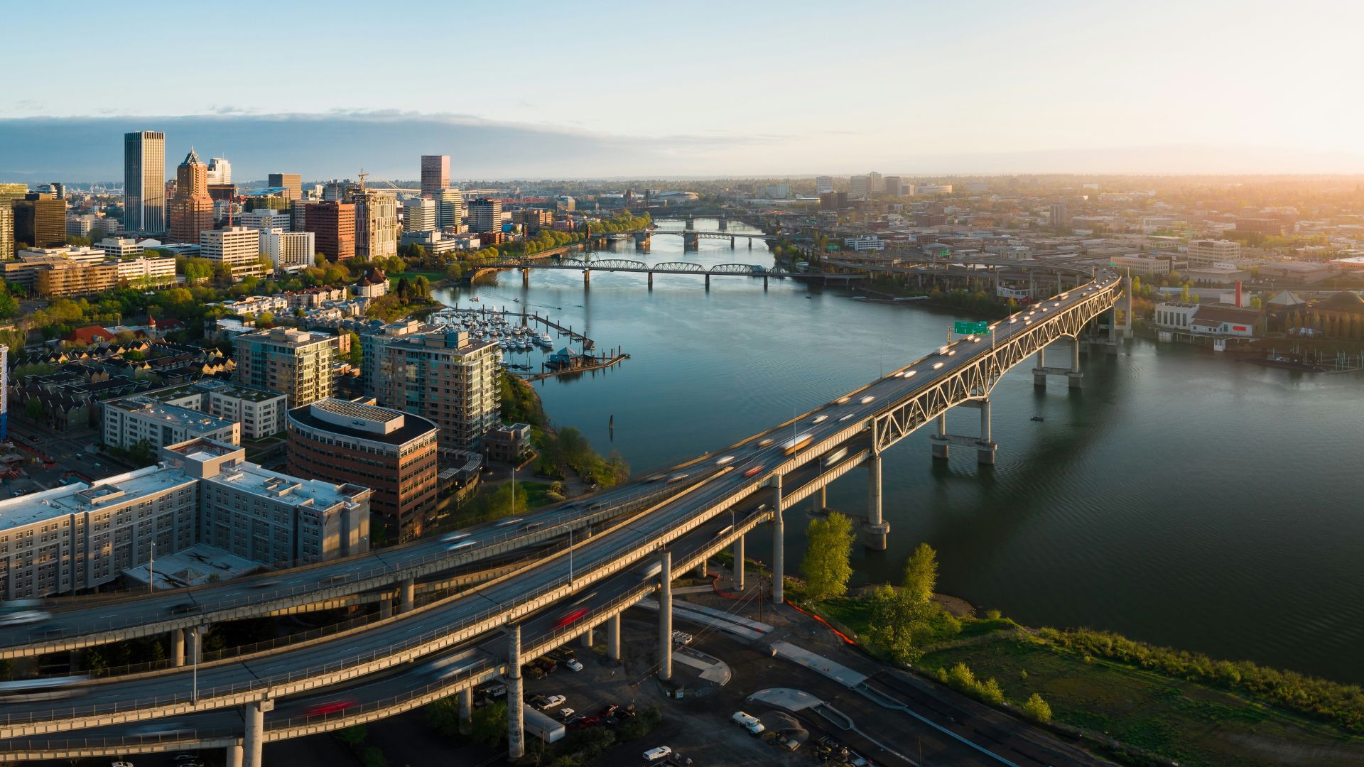 An aerial view of the Portland, Oregon downtown skyline and multiple bridges crossing the Willamette River, including the large Fremont Bridge and elevated highway ramps in the foreground, all under a golden evening light.