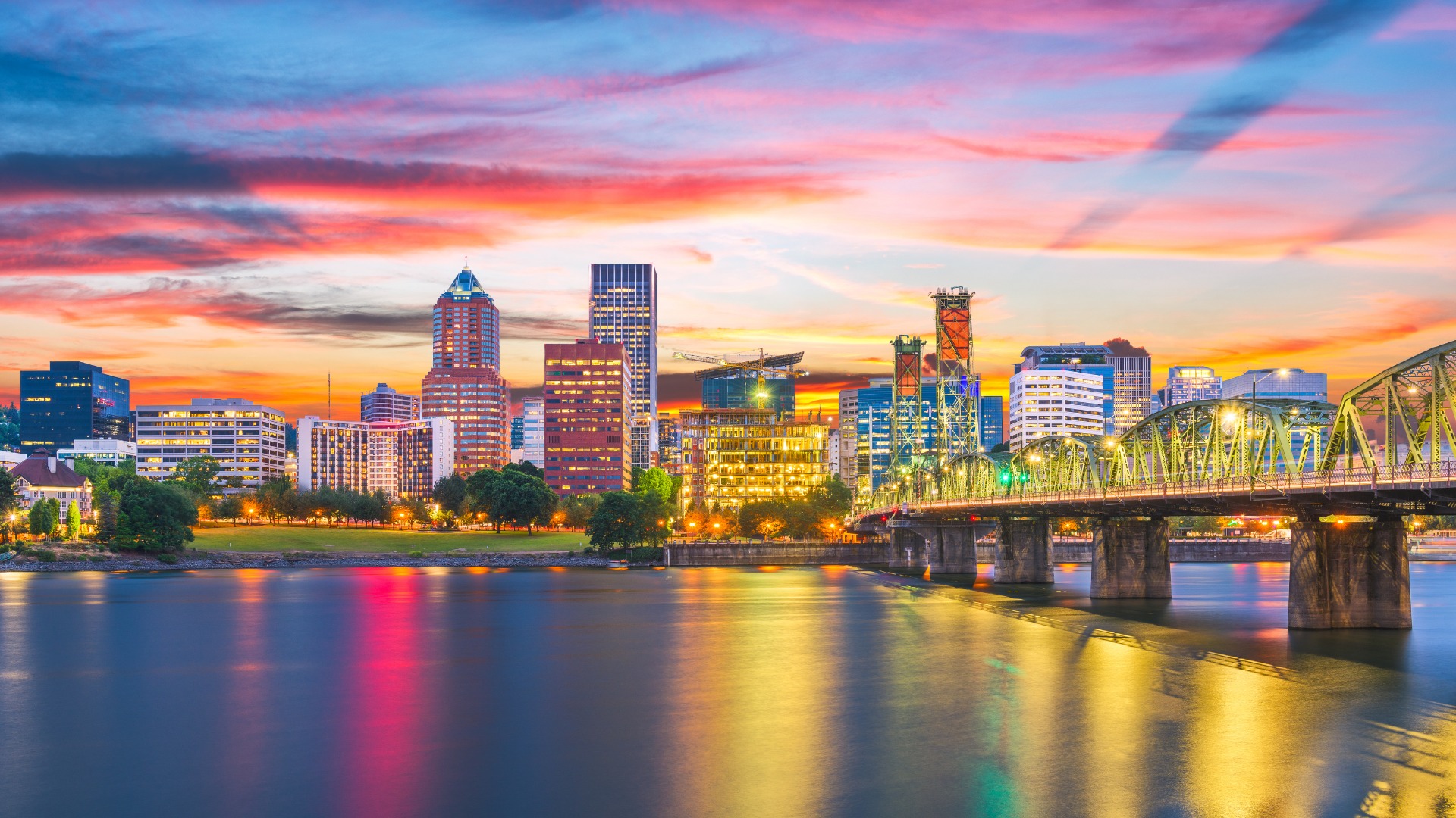A vibrant sunset view of the Portland, Oregon, downtown skyline, with various high-rise buildings illuminated against a colorful sky. The historic Hawthorne Bridge crosses the Willamette River in the foreground, with reflections visible on the water.