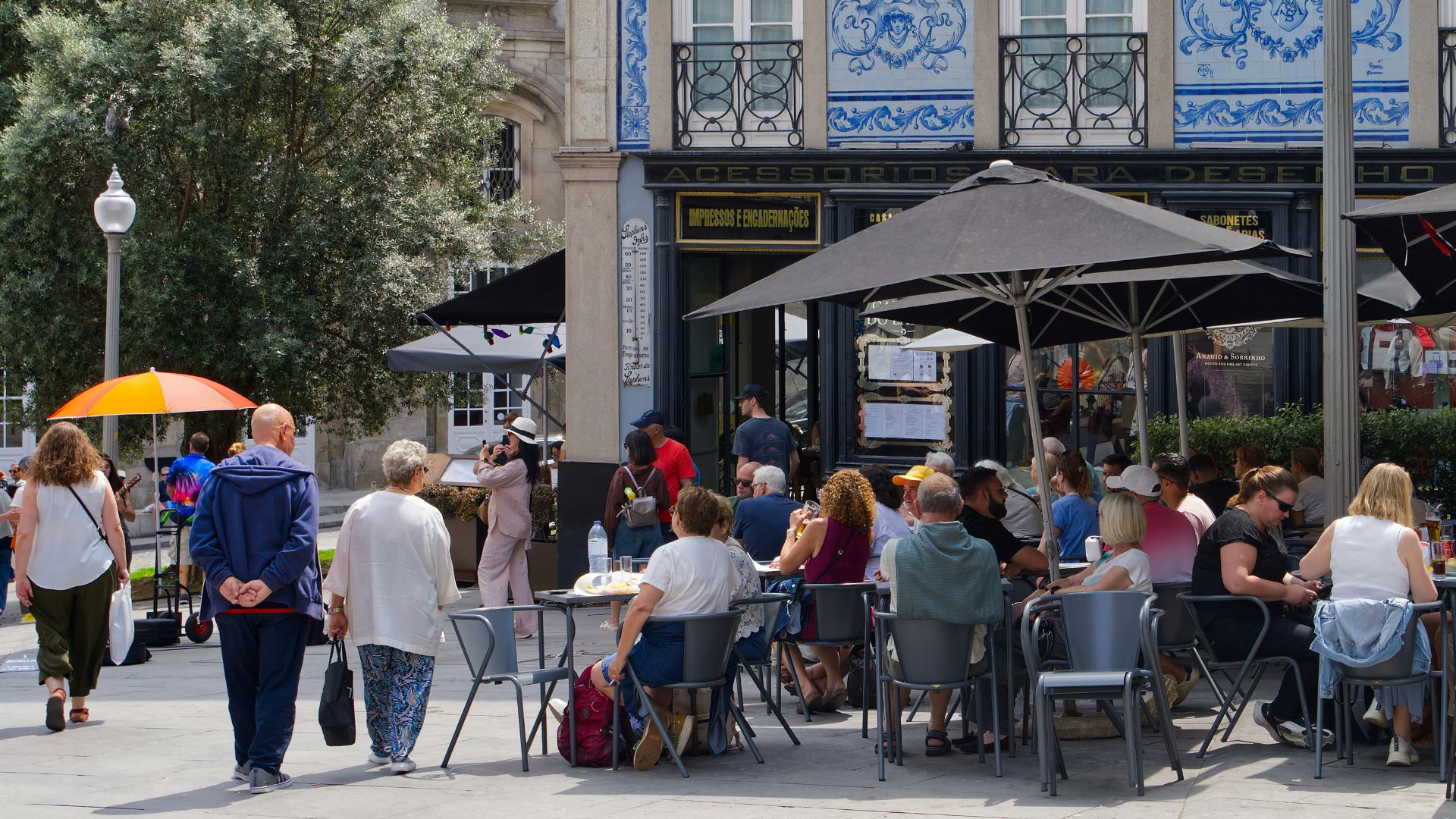 A vibrant daytime photograph of a busy pedestrian street in Porto, Portugal, lined with traditional buildings, an outdoor cafe under black umbrellas, and a building facade decorated with blue and white azulejo tiles.