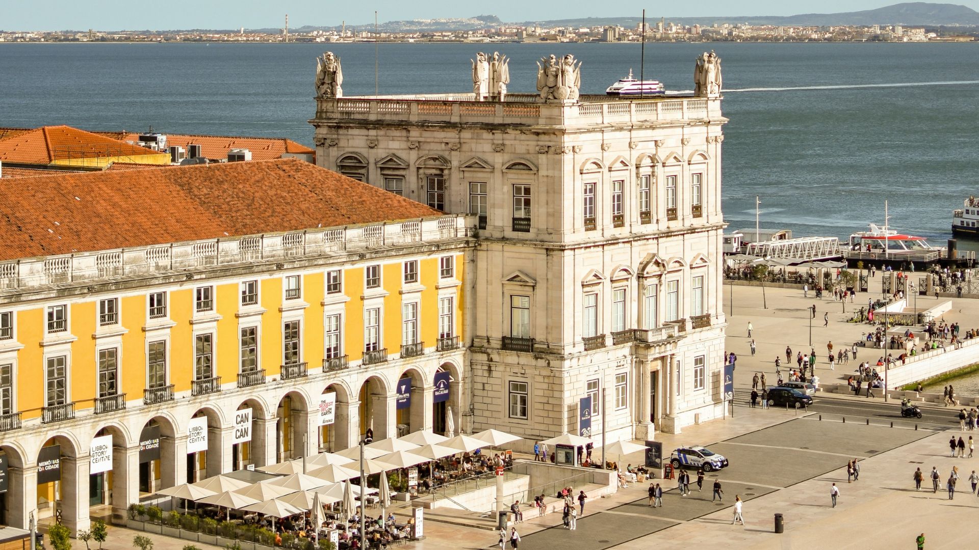 An elevated, sunlit photo of the waterfront Praça do Comércio in Lisbon, Portugal, showing historic yellow buildings with arcades, an open square with outdoor seating and pedestrians, and the broad Tagus River with distant hills in the background.