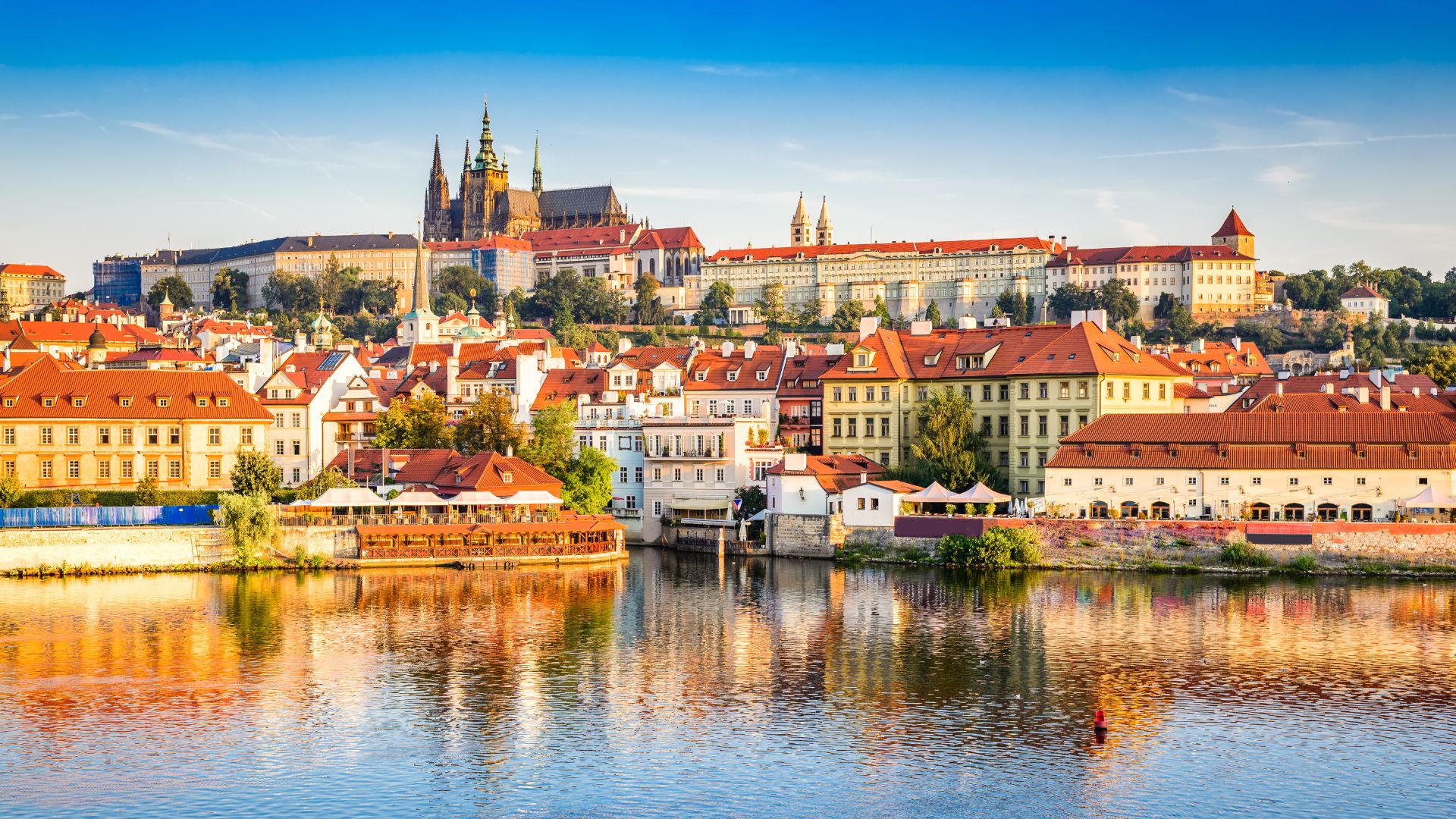 A scenic, daytime panoramic view of the historic Prague skyline, featuring the grand Prague Castle and the Gothic spires of St. Vitus Cathedral on a hill, with charming red-roofed buildings lining the banks of the Vltava River.