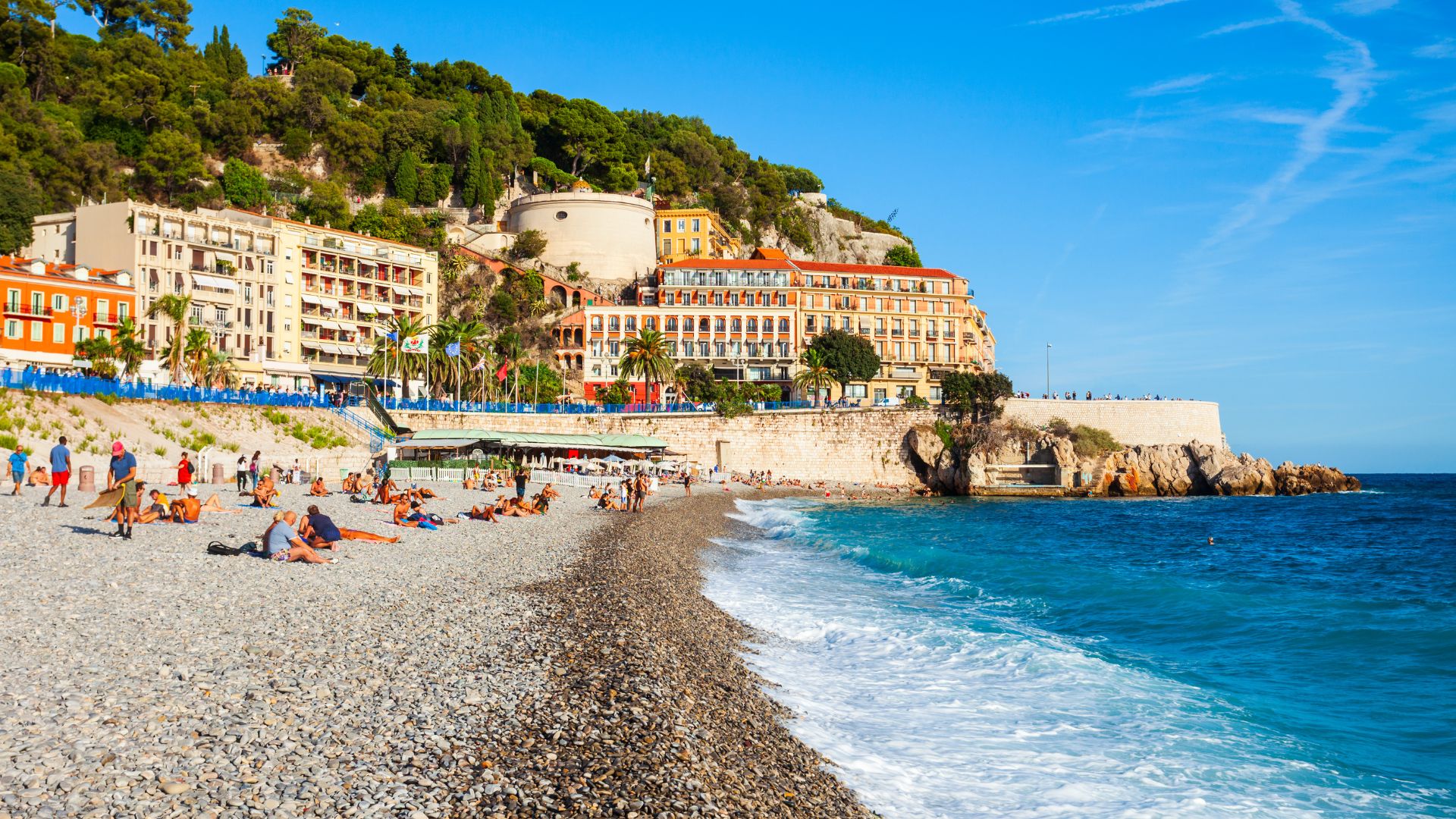 A sun-drenched, wide view of a vibrant pebble beach and the turquoise Mediterranean Sea in Nice, France, with a historic, fortified hillside and multi-story Belle Époque style buildings in the background.