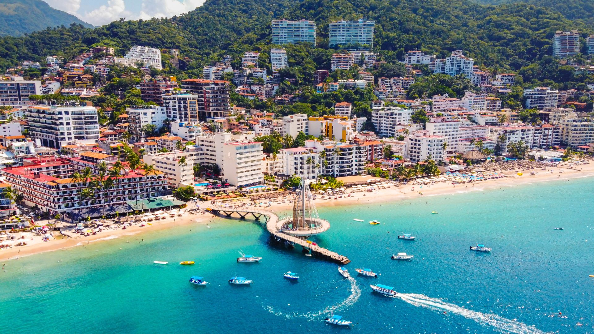 An aerial view of the cityscape of Puerto Vallarta, Mexico, with numerous multi-story buildings and hotels nestled into green, forested hillsides. In the foreground, a modern pier with a large, sail-shaped central structure extends into the vibrant turquoise ocean water, with several small boats nearby.