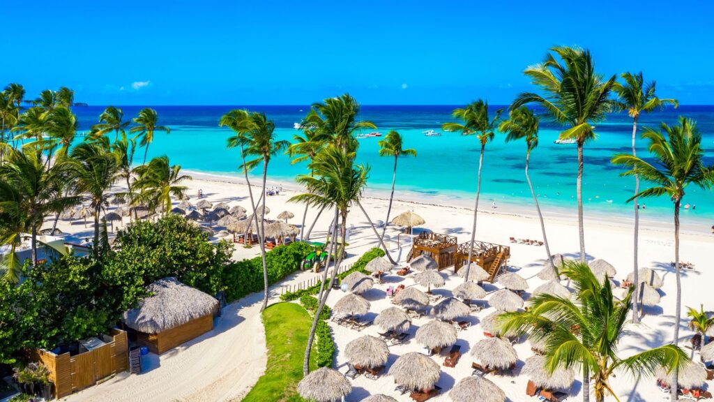 An aerial view of a tropical resort beach in Punta Cana, Dominican Republic, featuring vibrant turquoise ocean water, white sand, numerous thatched palapa umbrellas and huts, and lush green palm trees under a bright blue sky.