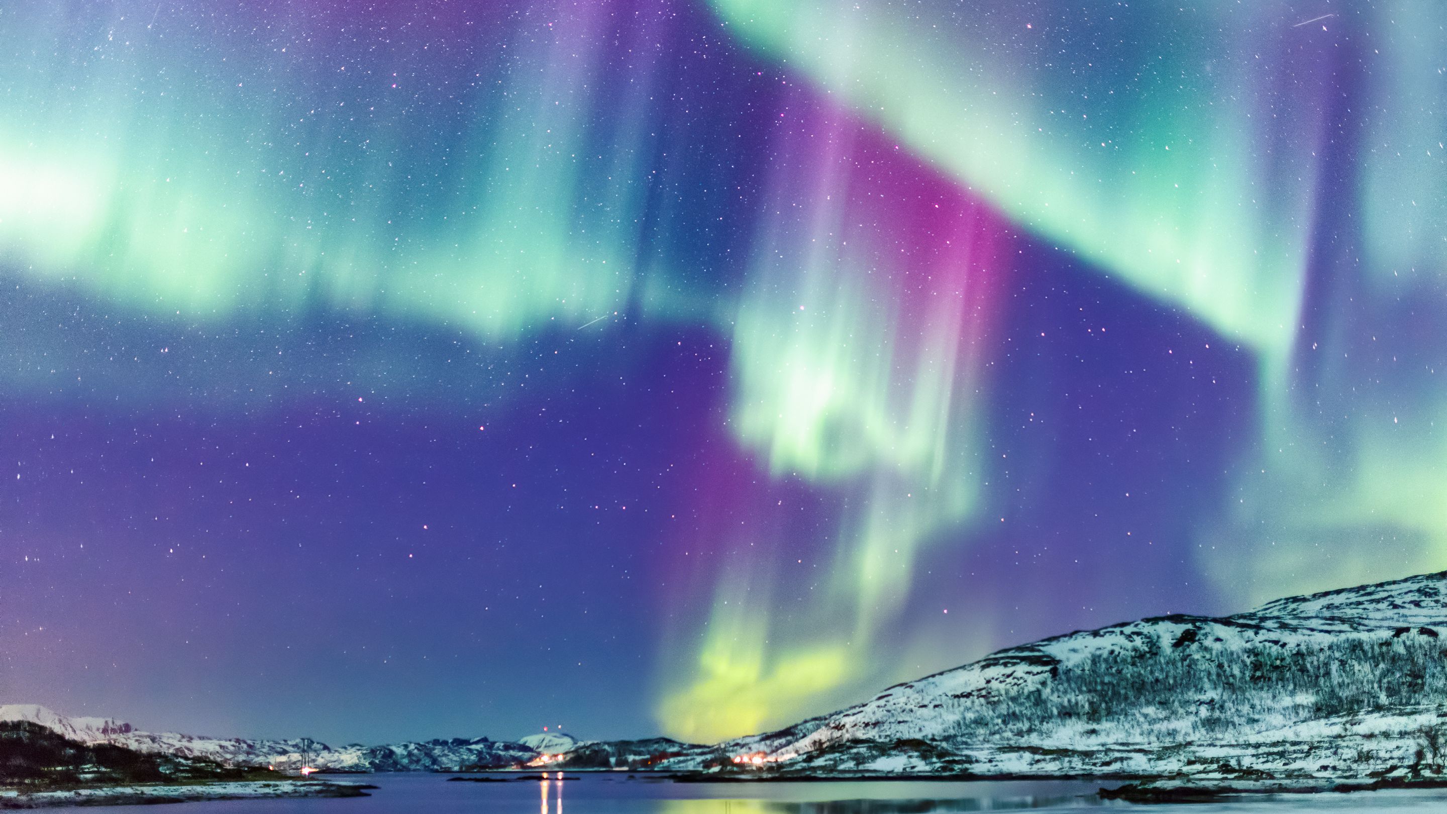 A night-time view of vibrant green and pink Aurora Borealis dancing across the sky above snow-covered hills and reflective water.