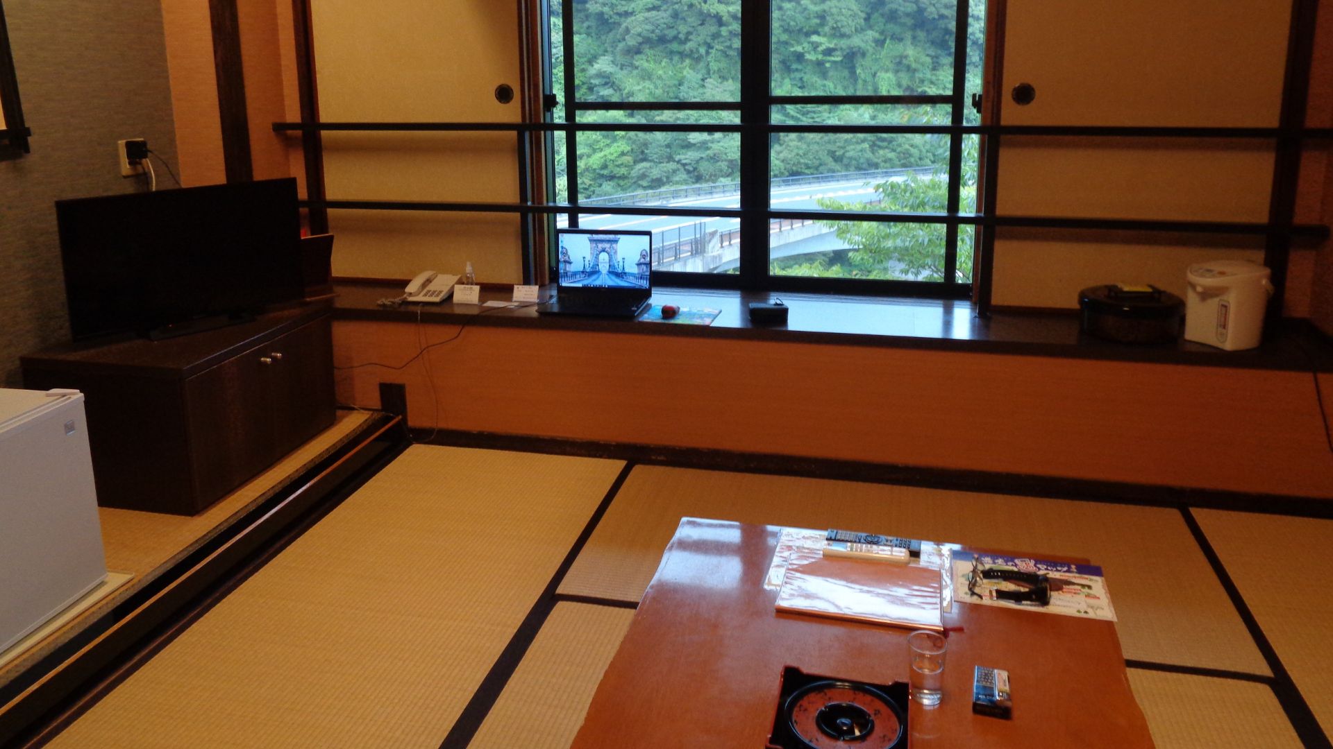 A traditional Japanese ryokan room with tatami flooring, shoji screens, a low wooden table with tea-making facilities, and a window view of a green, wooded valley and a bridge.