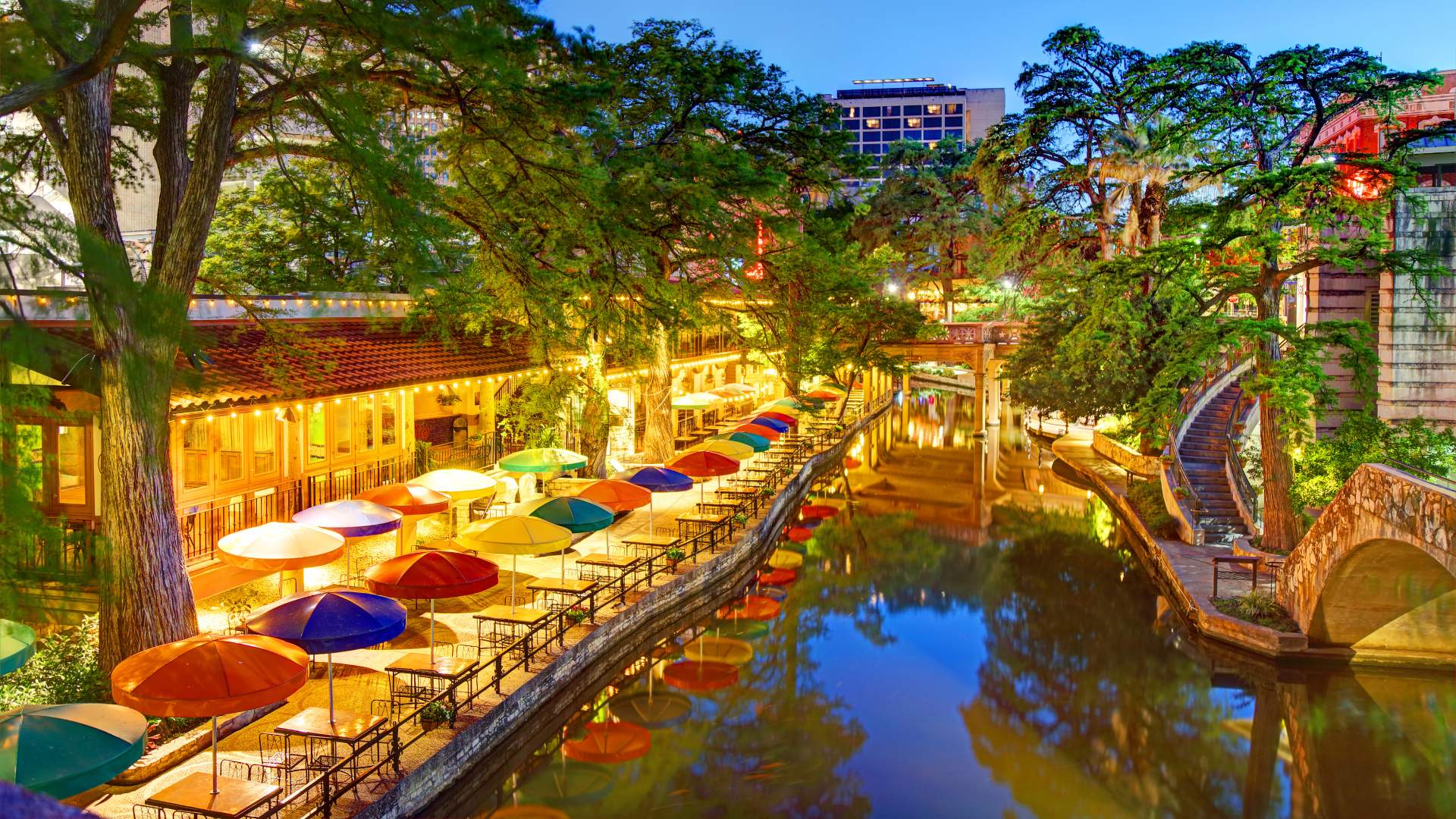 A scenic evening view of the San Antonio River Walk in Texas, featuring outdoor dining patios with vibrant multi-colored umbrellas reflected in the tranquil water, lined by lush bald cypress trees and illuminated by warm lights.